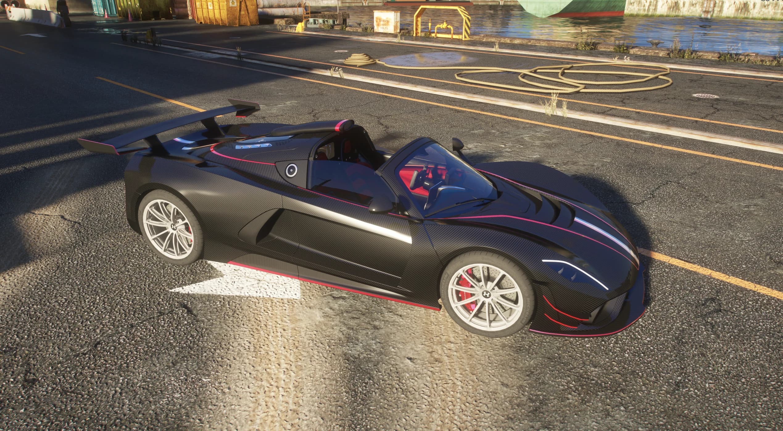 High-angle view of the black carbon Venom F5 Roadster with its open cockpit, red interior and swan-neck rear wing, on a dock