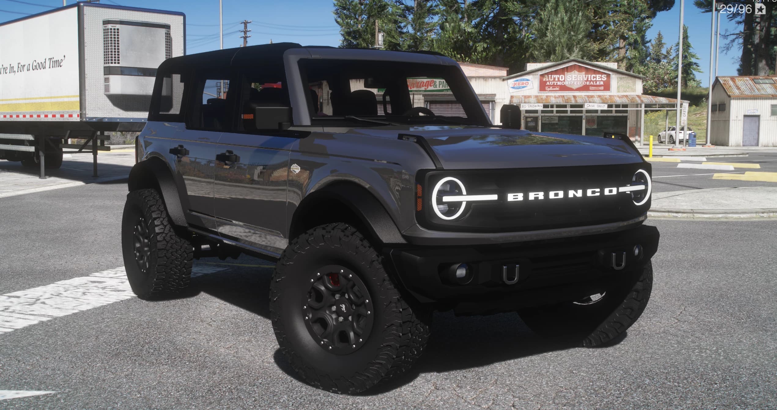 Grey four-door Ford Bronco with an illuminated BRONCO grille, black top and off-road tyres, front three-quarter view at a gas station