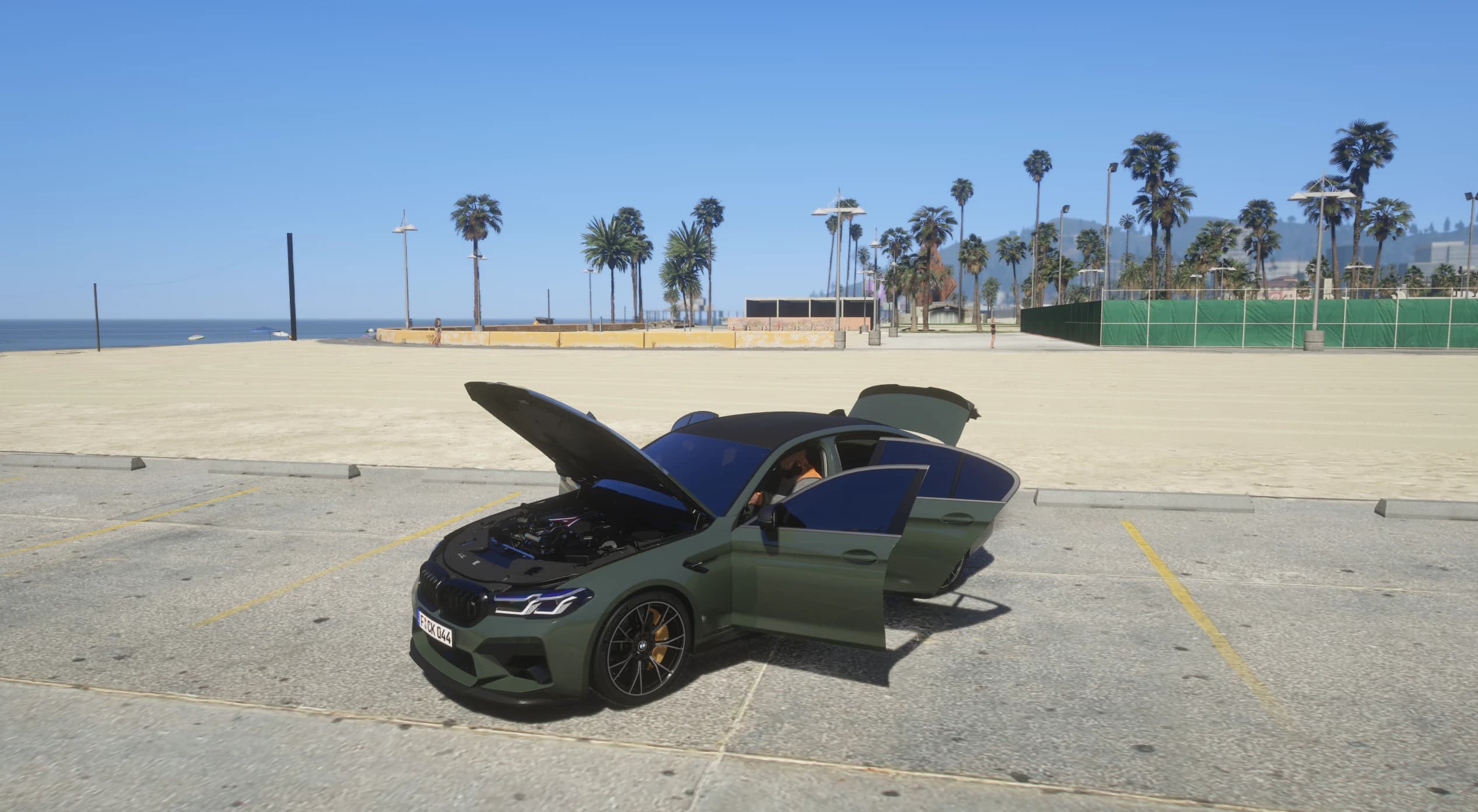 The M5 with its hood and driver's door open, revealing the blue-accented engine bay and a player character sitting inside, parked by the beach with palm trees and the ocean behind