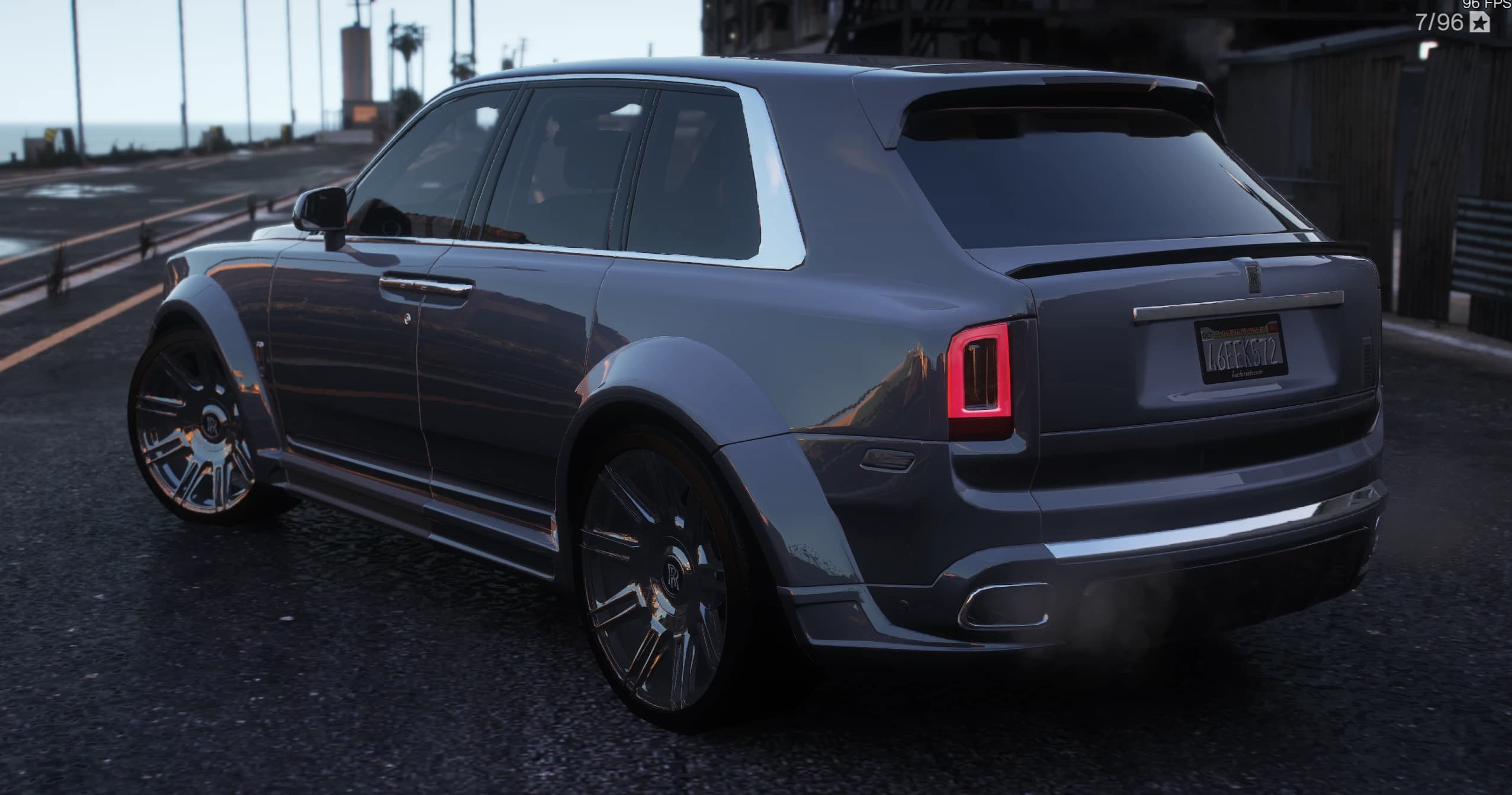 Rear three-quarter of a gunmetal grey Cullinan on a dockside road at sunset, showing the roof spoiler, the lip on the tailgate, the vertical red tail lights and the four exhaust tips