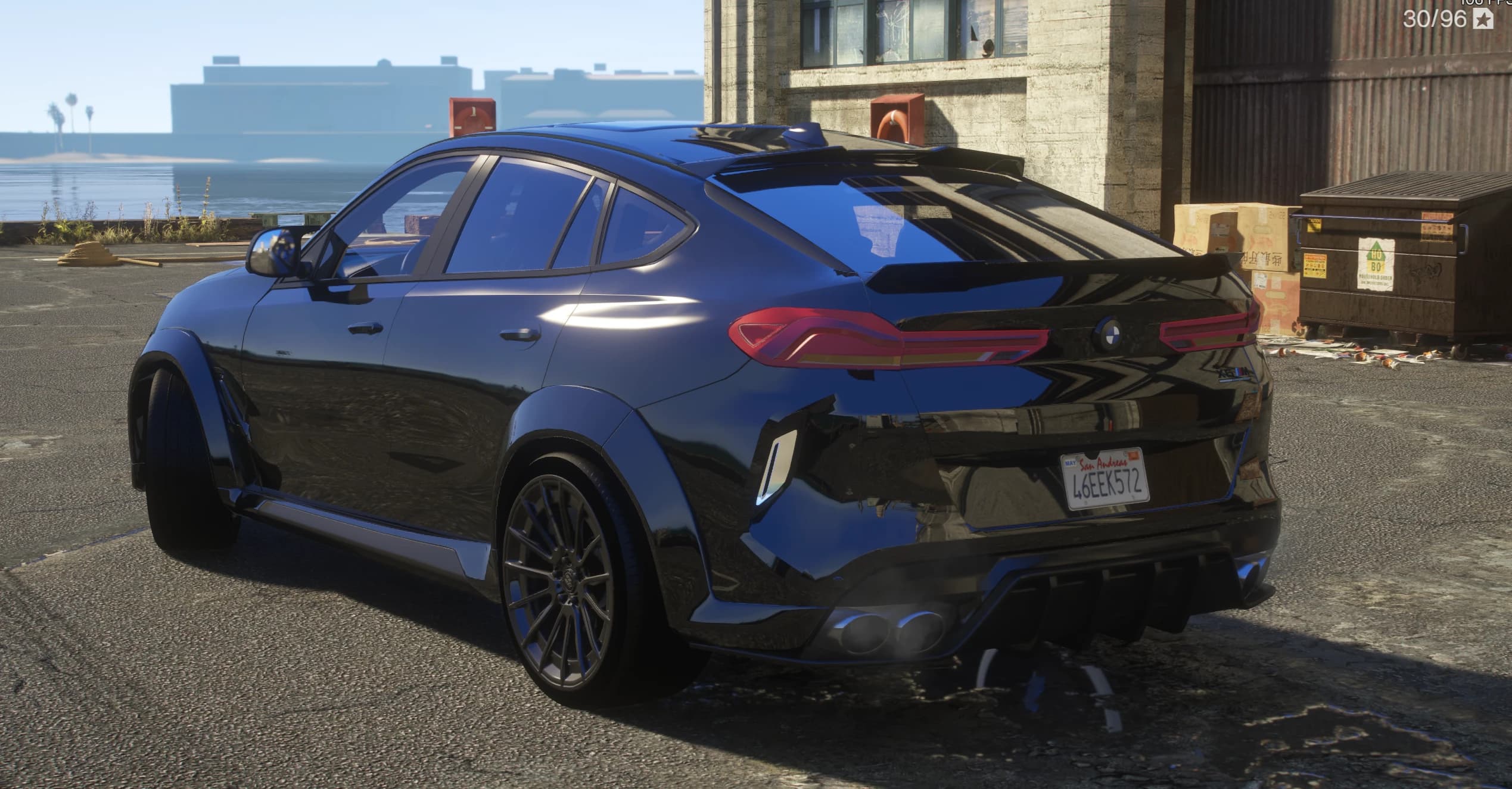 Rear three-quarter of the X6M at a dock, showing the widebody flares, the ducktail spoiler, the full-width connected tail lights, the BMW roundel, quad exhausts and a San Andreas plate reading 46EEK572
