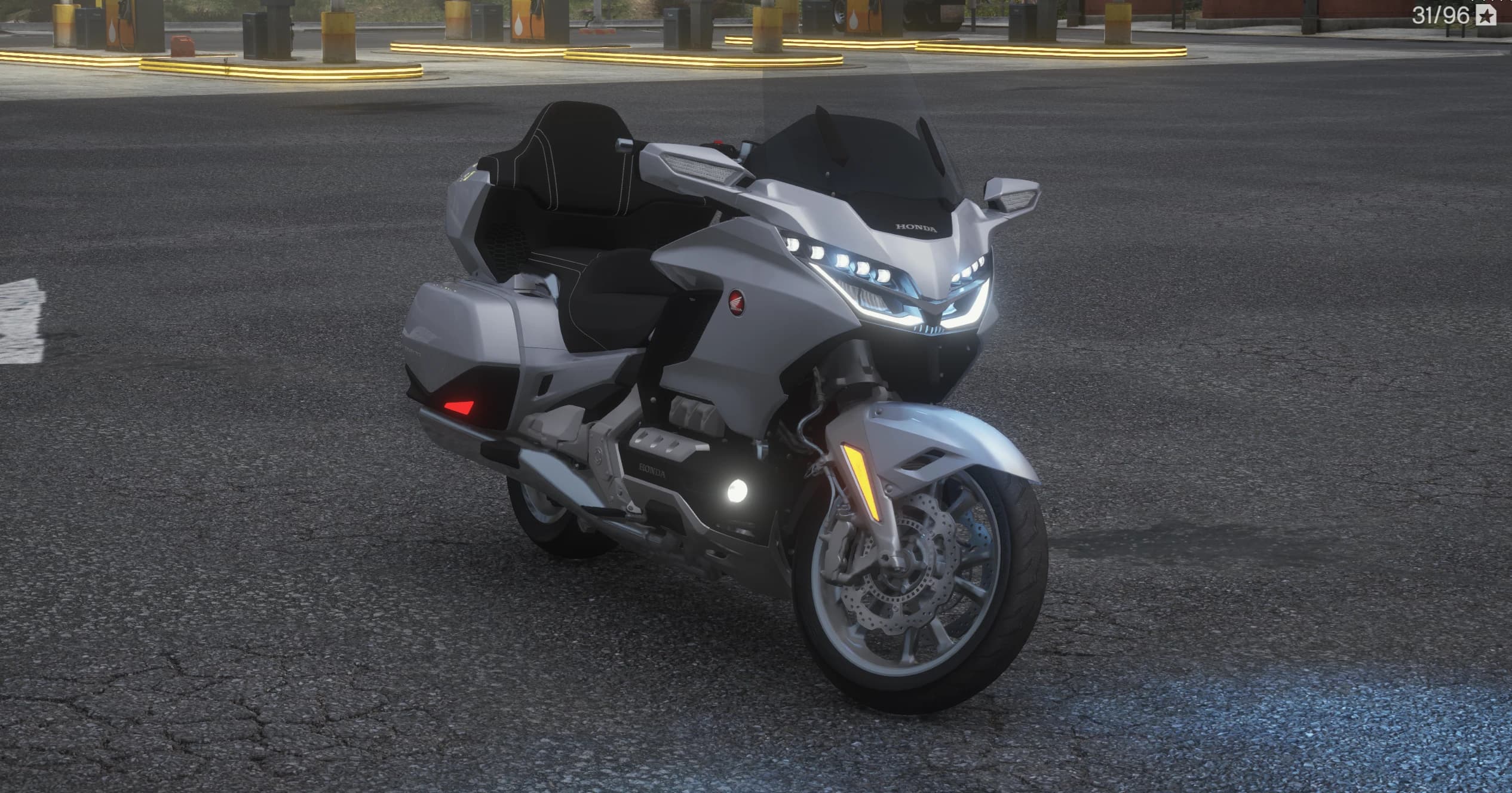 Front three quarter view of the silver Honda Goldwing Touring at a gas station with the split LED headlights lit, tall passenger backrest and side panniers