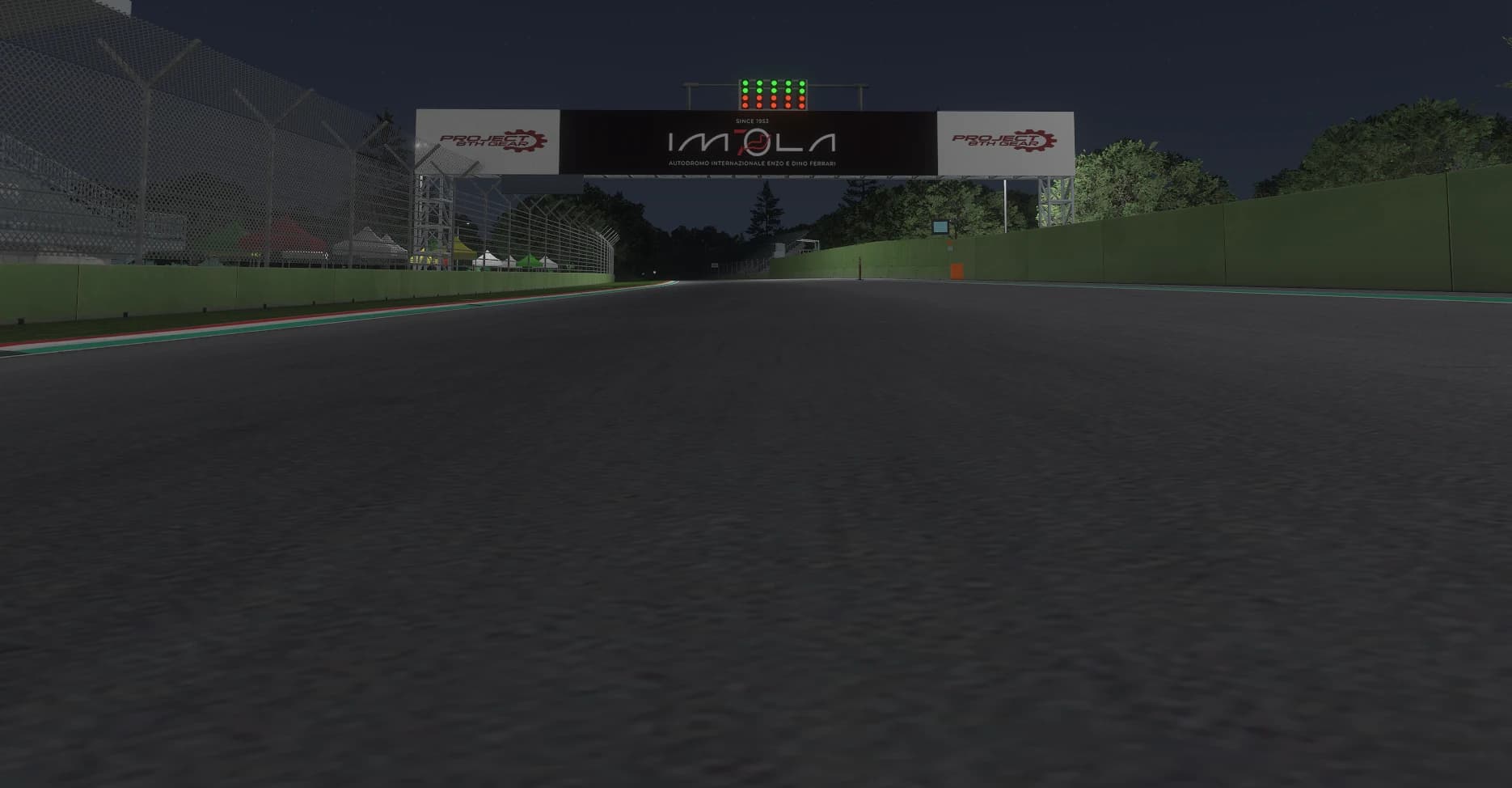 Night view from the Imola start line looking up the straight: the lit IMOLA gantry reading Autodromo Internazionale Enzo e Dino Ferrari, the start lights above it, the grandstand on the left and the green trackside walls