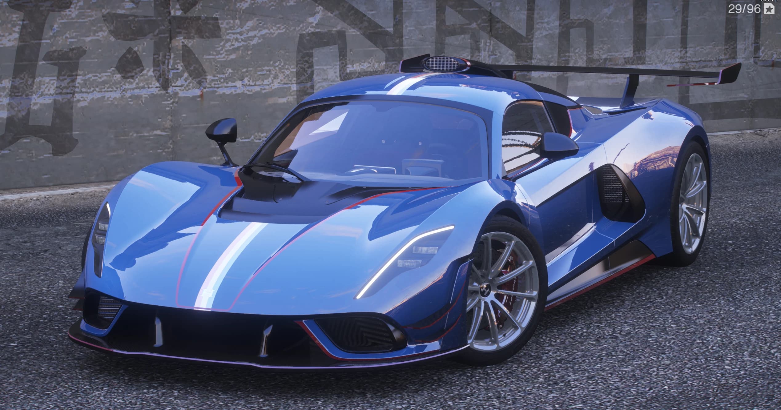 Blue Hennessey Venom F5 Revolution Roadster hypercar with a white-and-red centre stripe and a large rear wing, front three-quarter view against a graffiti wall