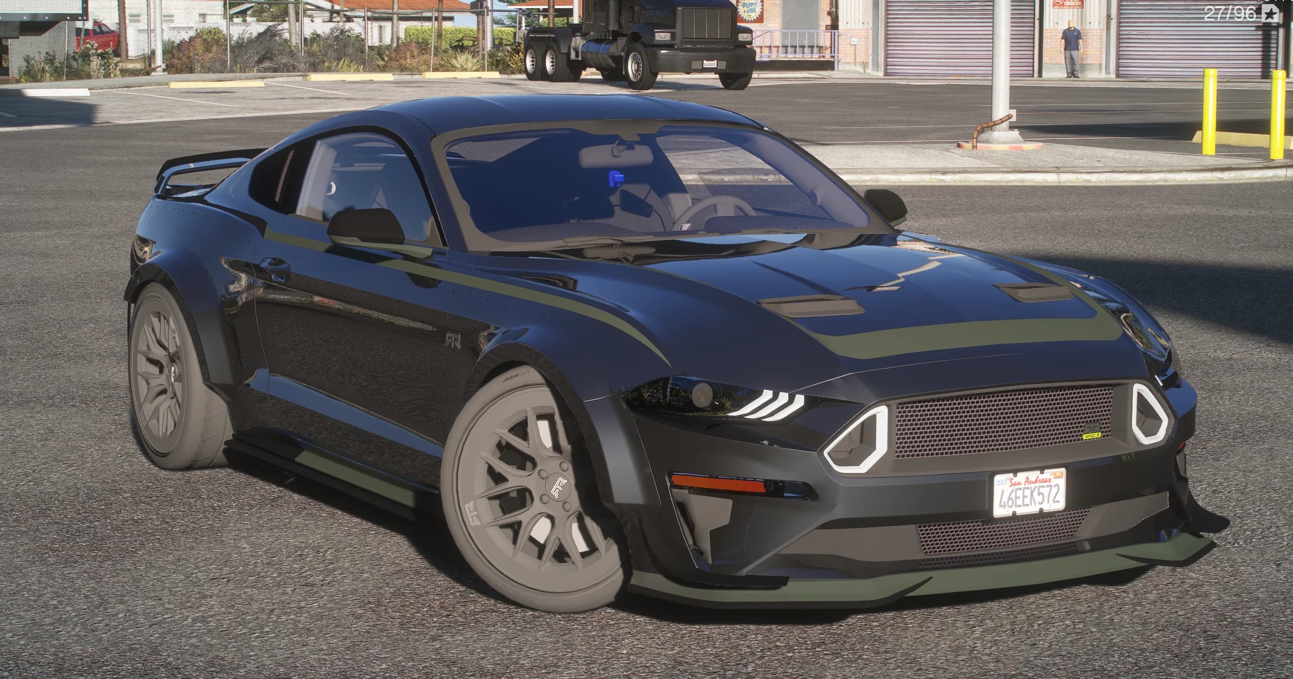 Black 2015 Ford Mustang RTR Spec 5 in an industrial yard, front three-quarter view with olive green stripes over the hood, bolted-on fender flares, a deep front splitter and grey multi-spoke wheels