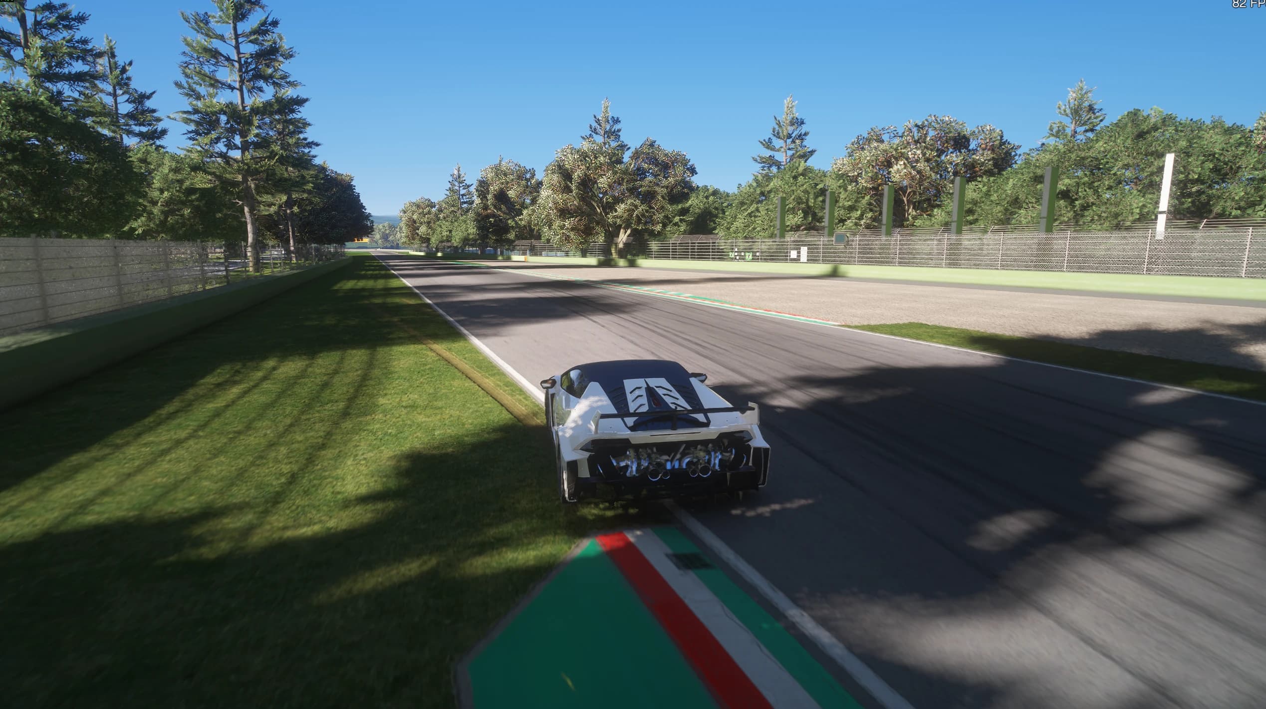 Daytime chase-cam shot of a white supercar on a tree-lined section of the track, with grass verges, a gravel trap on the right and red and white kerbs