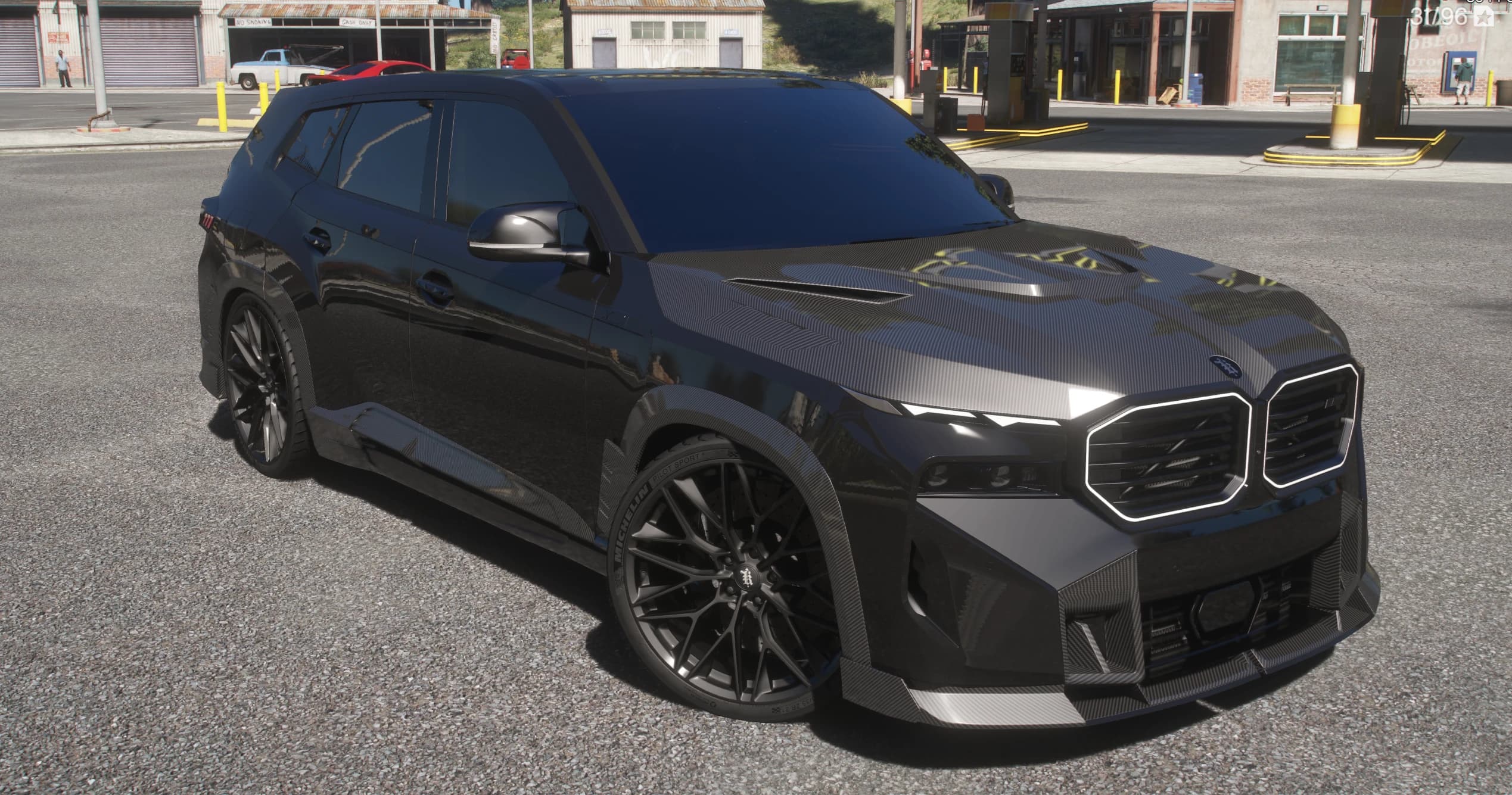 Front three quarter view of the black BMW XM Larte at a rural gas station, with the carbon hood, lit kidney grille outline and Michelin tyres