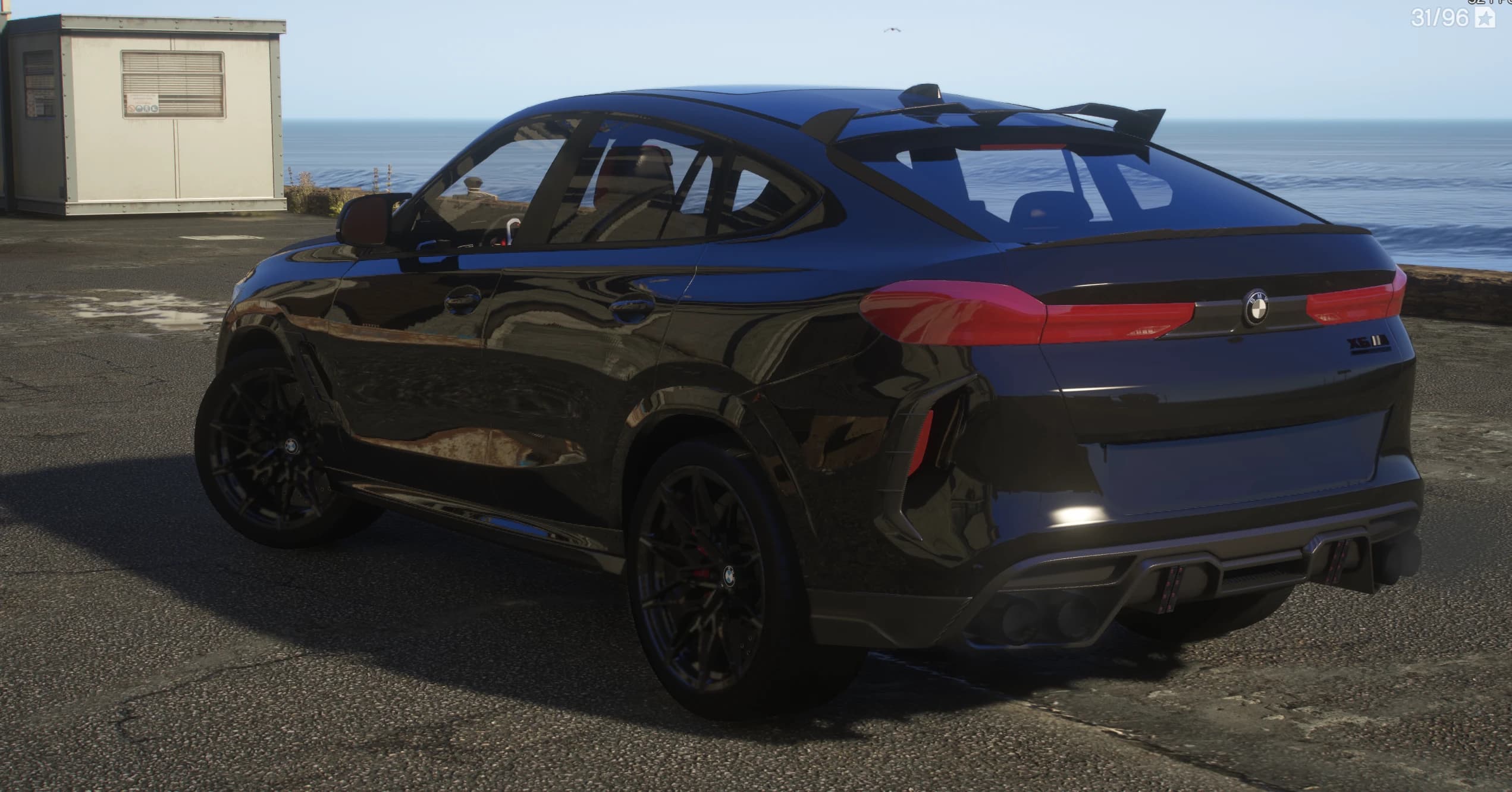 Rear three quarter view by the sea showing the roof spoiler with side winglets, the X6 M badge, quad exhaust tips and black mesh wheels