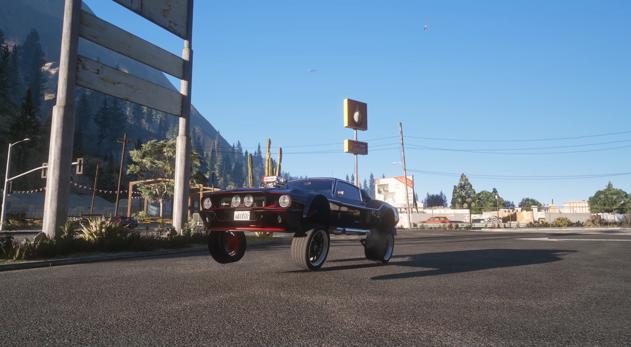 Low camera angle on a rural road: the Shelby has its nose hoisted on the hydraulic suspension with both front wheels hanging clear of the asphalt, next to a Fast Serve Gasoline sign