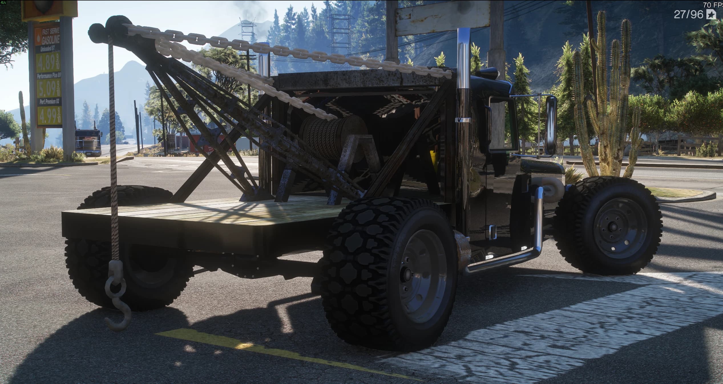 Rear three-quarter of the Tow-Rod parked at a petrol station, showing the wooden flatbed deck, the welded steel boom with its chain and hook and the winch drum with its wound cable