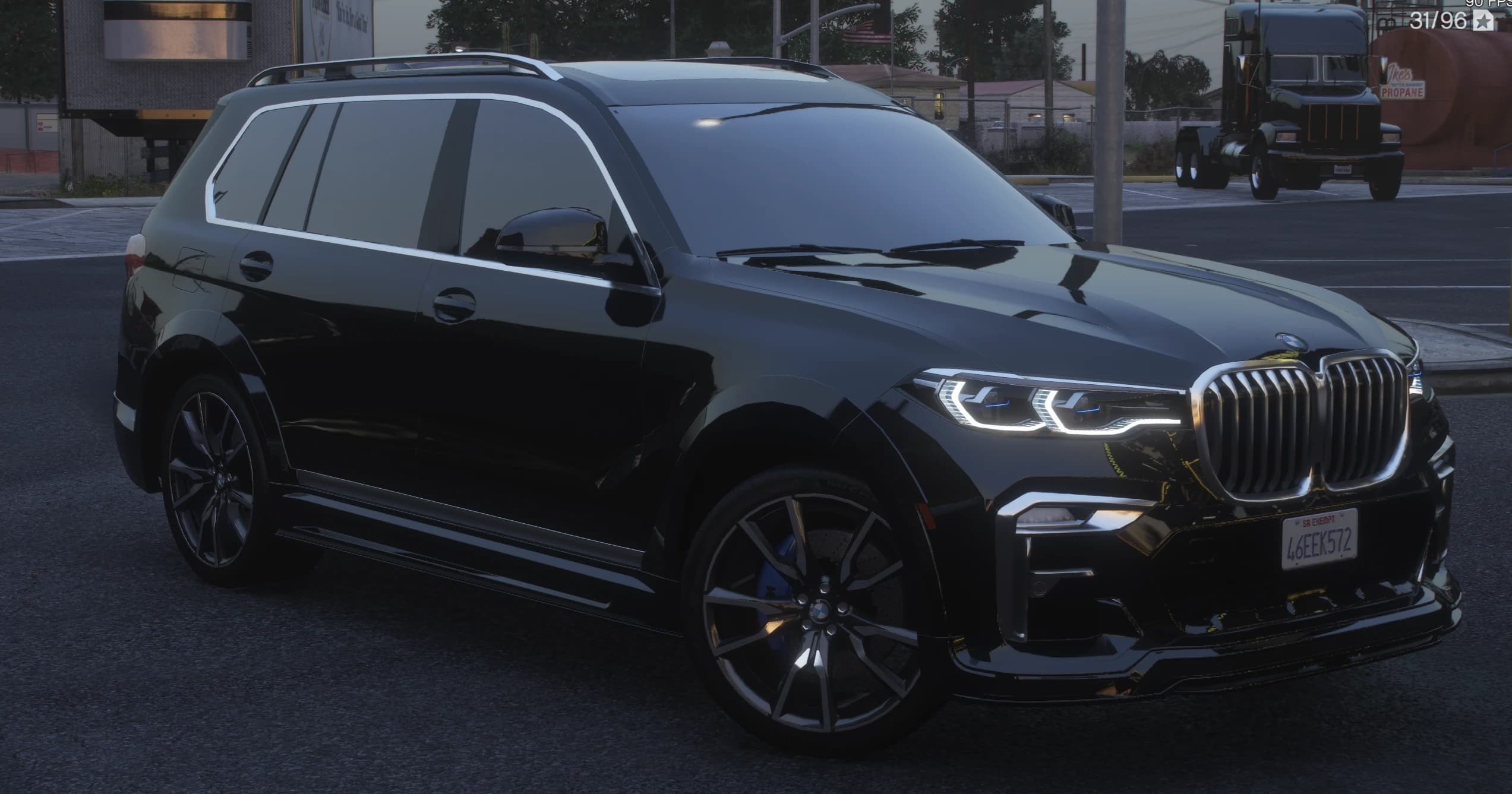 Front three quarter view of the black X7 at dusk with the big illuminated kidney grille, lit LED headlights and blue M calipers behind multi spoke wheels