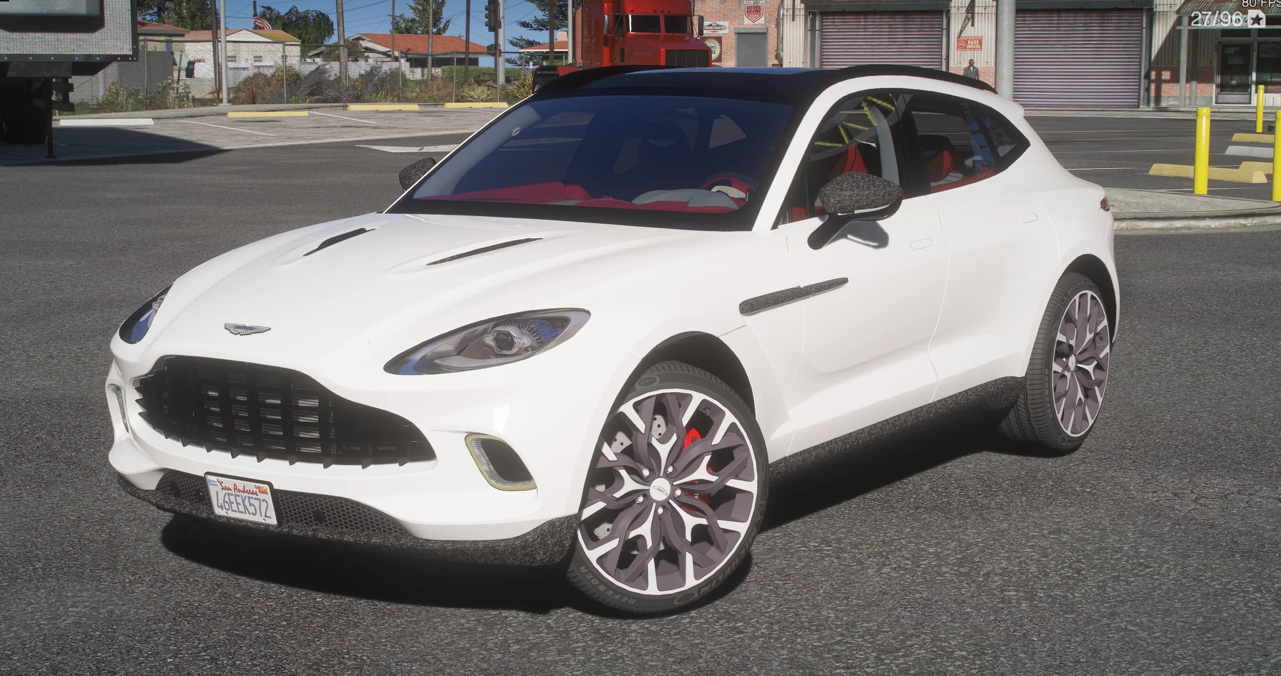 White 2025 Aston Martin DBX Carbon Edition in an industrial yard, front three-quarter view with the black roof, the carbon-look mirror caps and side vents, grey multi-spoke wheels and red brake calipers