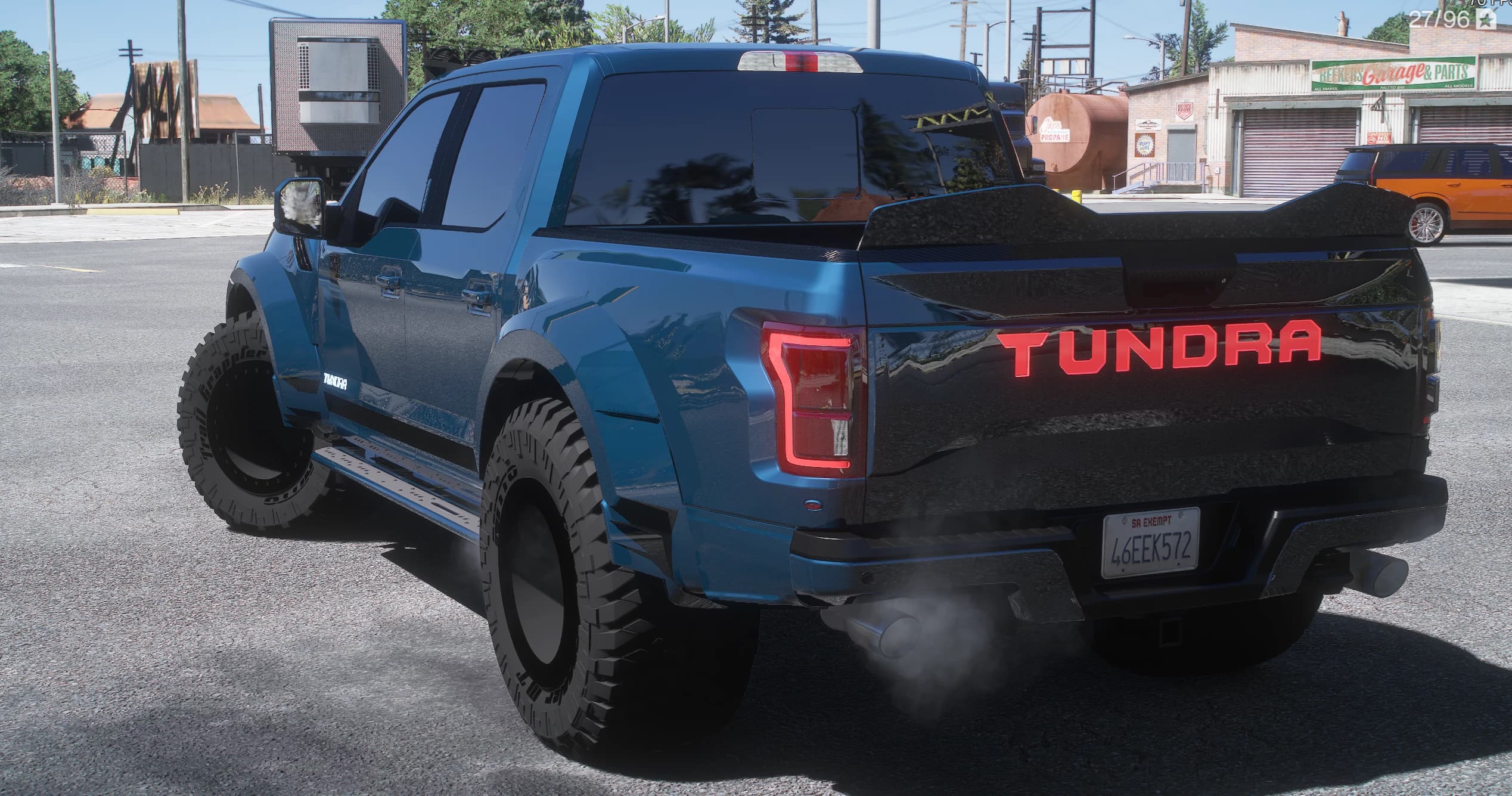 Rear three-quarter of the blue Tundra outside a garage, showing the illuminated red TUNDRA lettering across the tailgate, the black bed cover, the side steps and smoke from the exhaust