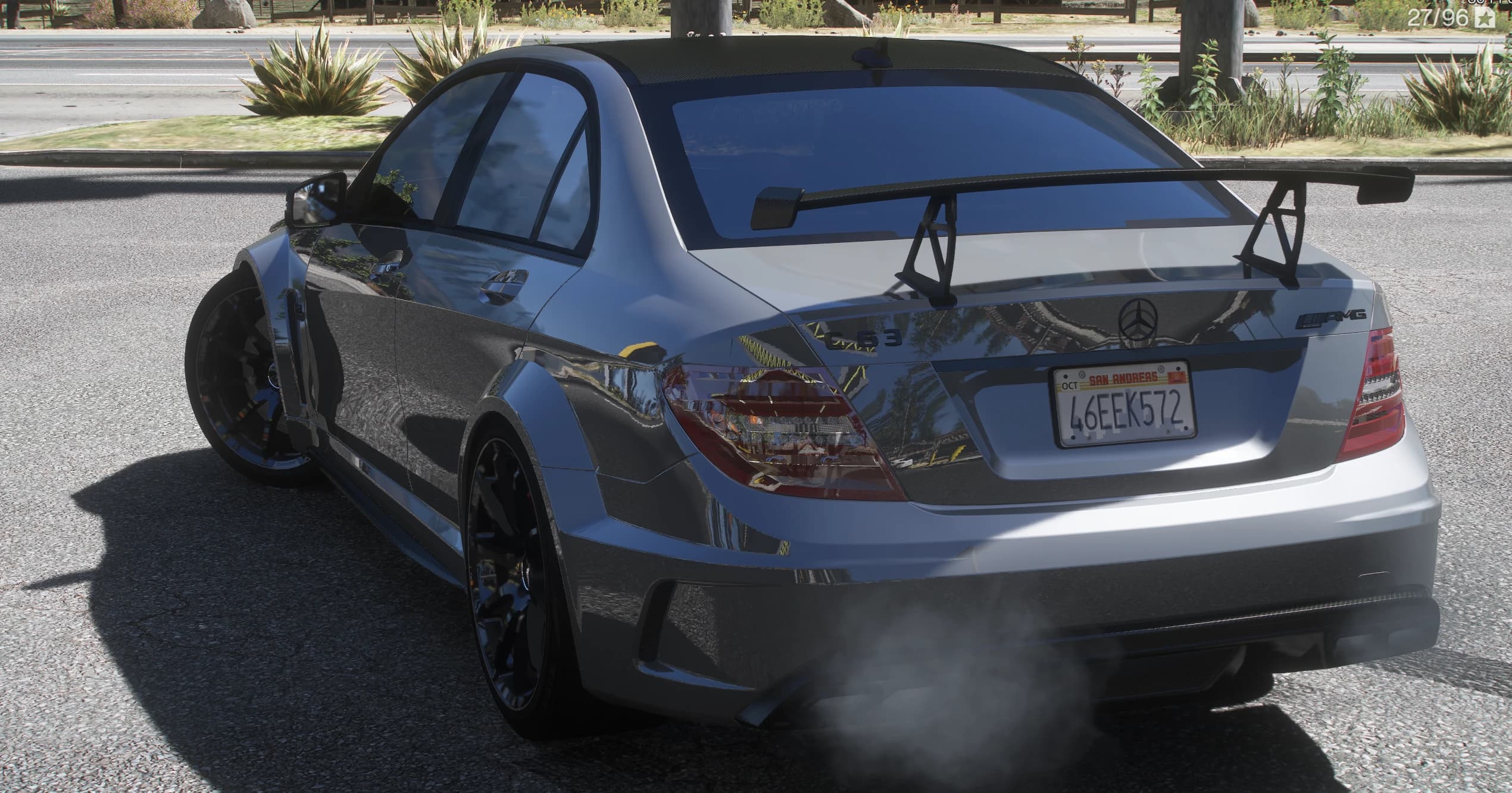 Rear three-quarter of the silver C63 at the roadside, showing the big black GT wing on its uprights, the C 63 and AMG badges, the carbon roof and smoke from the quad exhaust