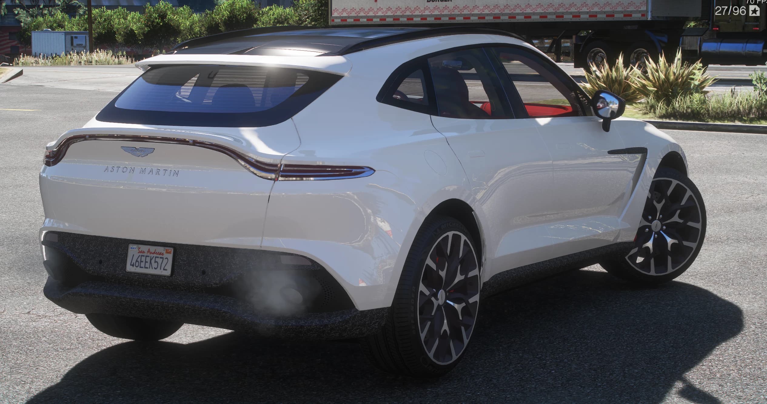 Rear three-quarter of the white DBX with the ASTON MARTIN lettering across the tailgate, the roof spoiler, the red leather cabin visible through the glass and exhaust smoke under the diffuser
