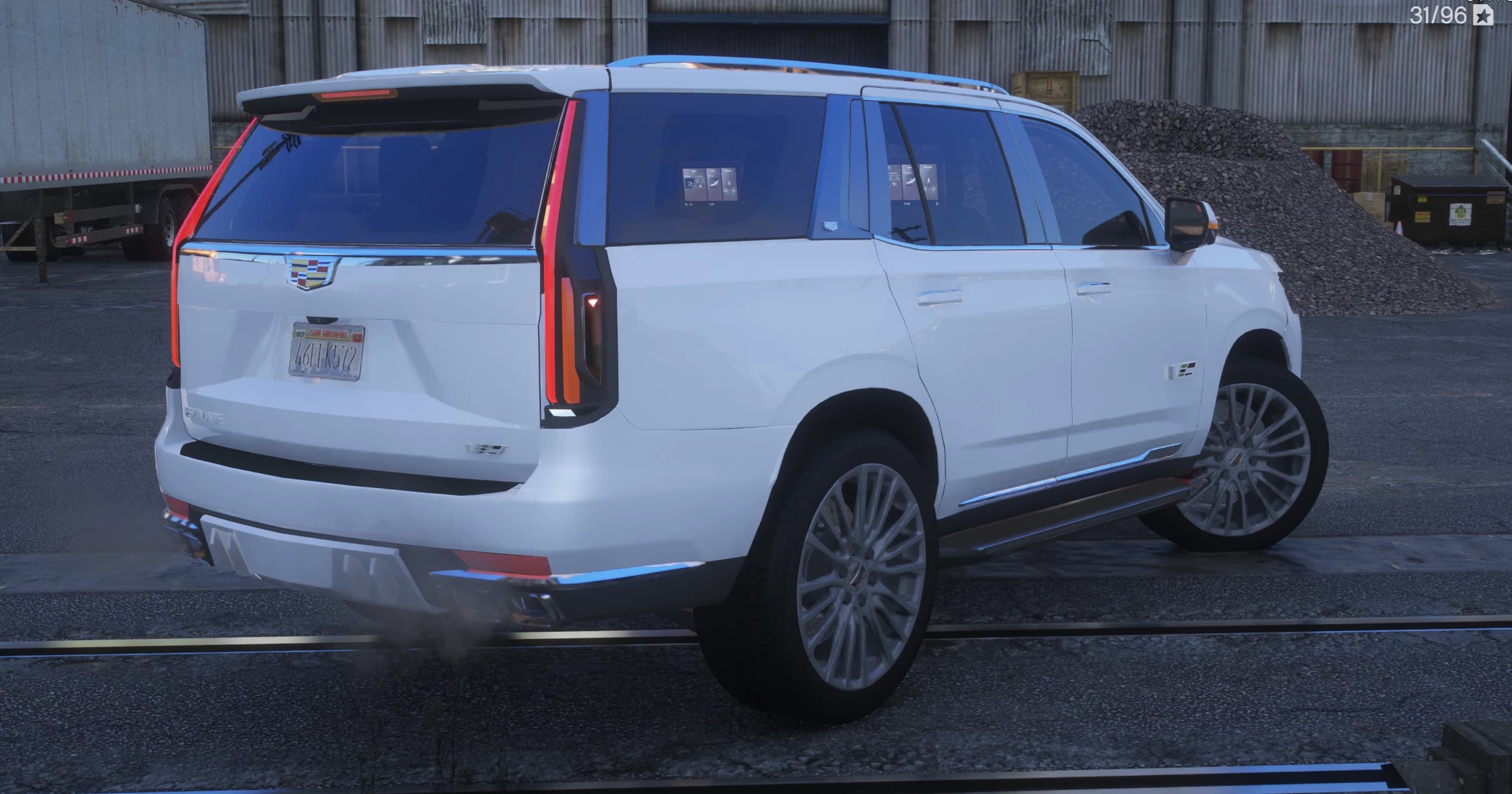 Rear three quarter view in an industrial yard showing the vertical taillights, the Cadillac crest on the tailgate, quad exhaust tips and the rear seat screens visible through the glass