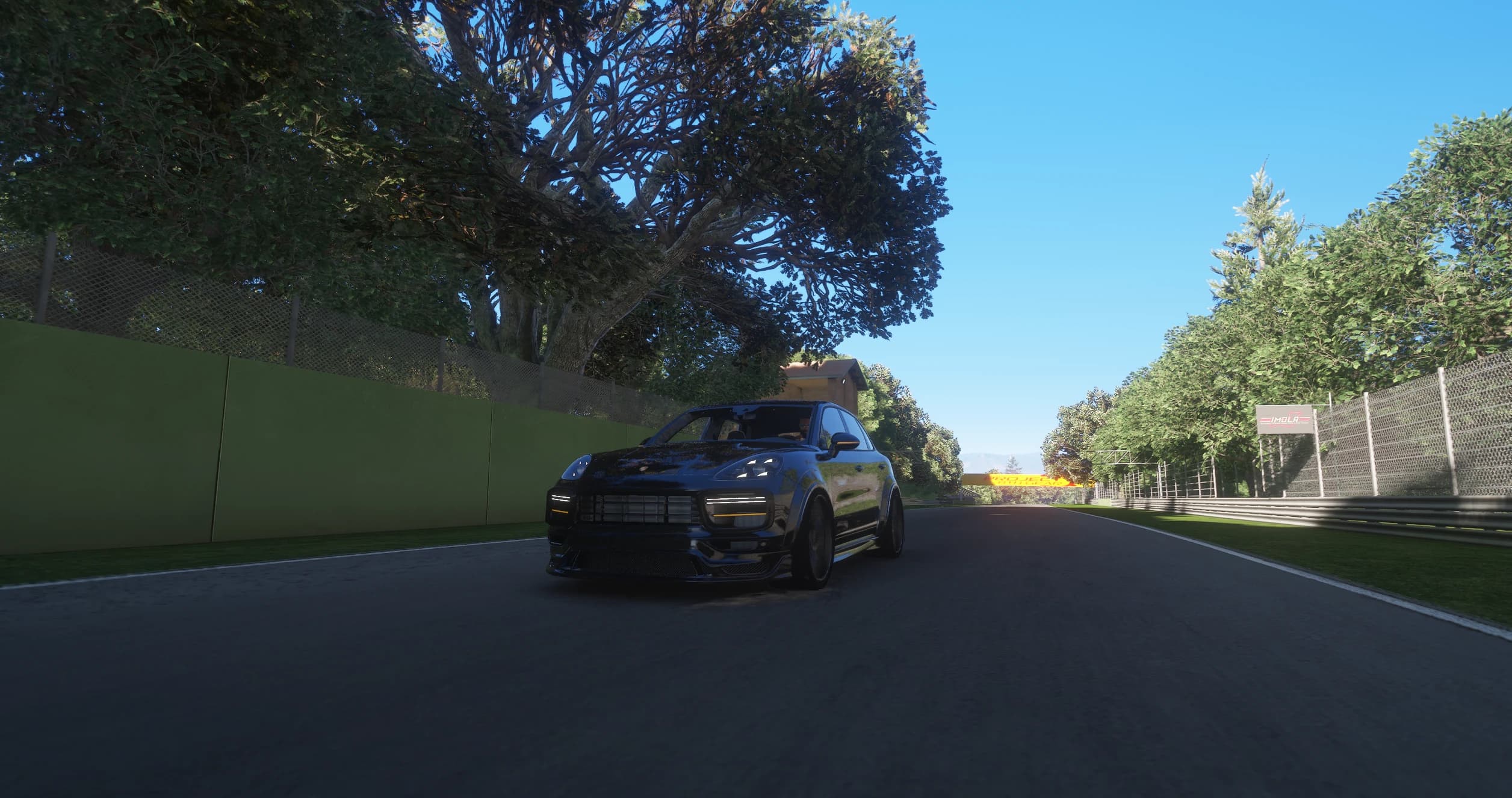 The Cayenne driving down a tree-lined stretch of the Imola circuit in the morning with the LED daytime lights and the amber front indicators switched on, an IMOLA board on the fence to the right