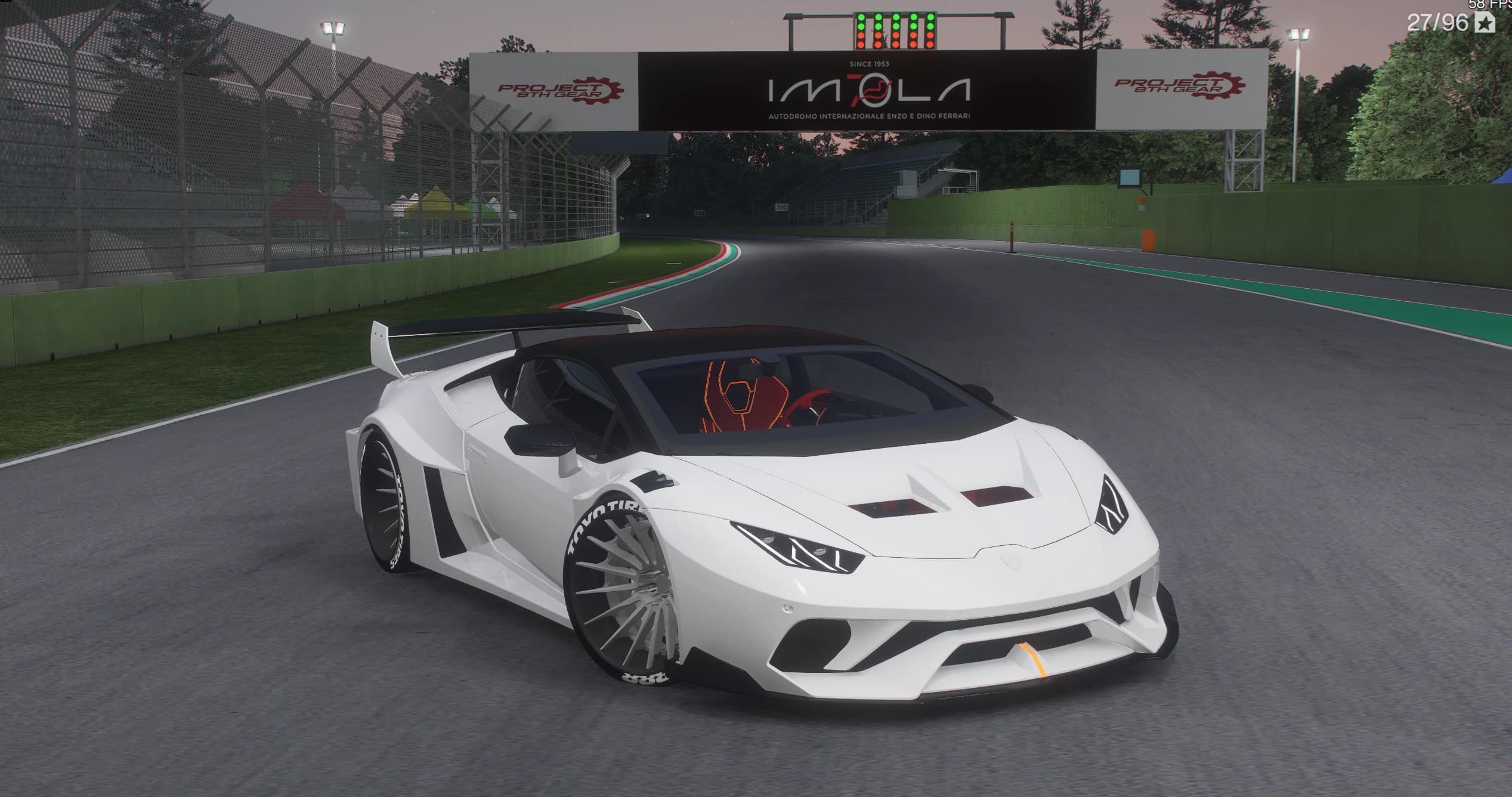 White Lamborghini Huracan with the Liberty Walk widebody kit parked on the Imola start straight at dusk, front three-quarter view under the IMOLA gantry, showing the bolted arches, the roll cage through the glass and the swan-neck rear wing