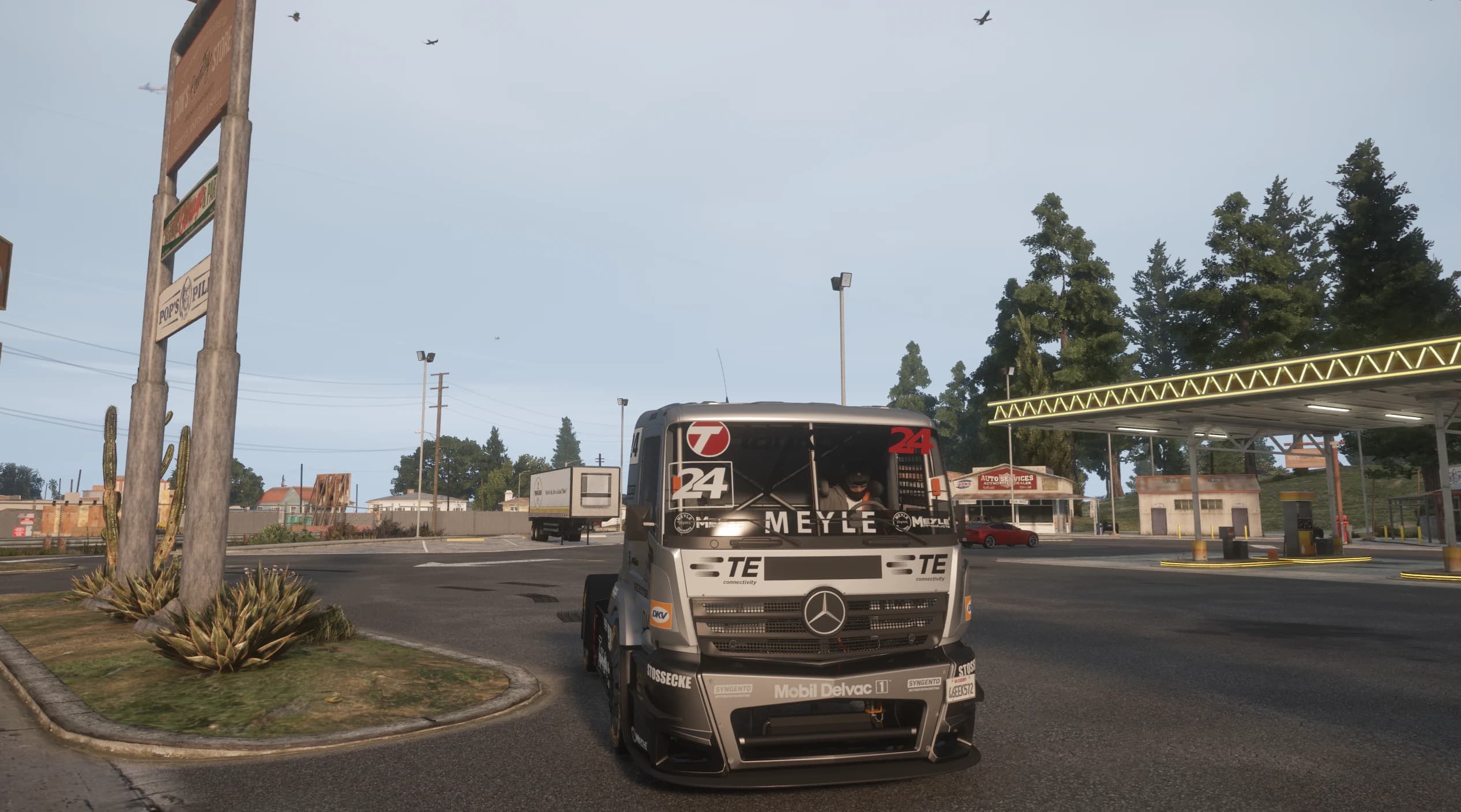 Head on view of the race truck rolling past a country gas station with cacti and a store sign, helmeted driver visible through the windscreen