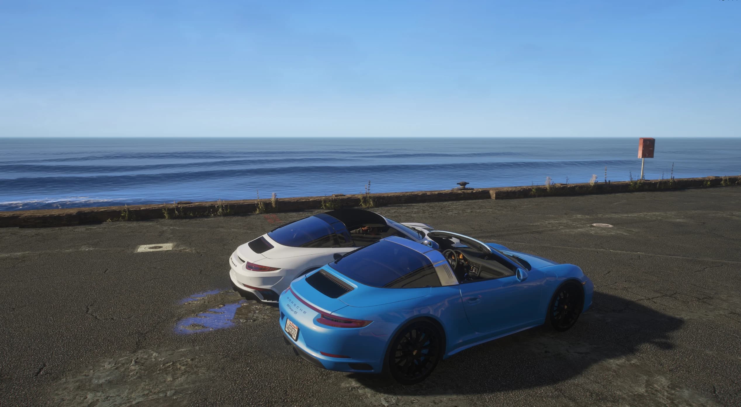 High-angle rear three-quarter of both cars parked beside the ocean seawall, with the blue Targa 4S and its silver Targa hoop in the foreground and the white Stinger behind