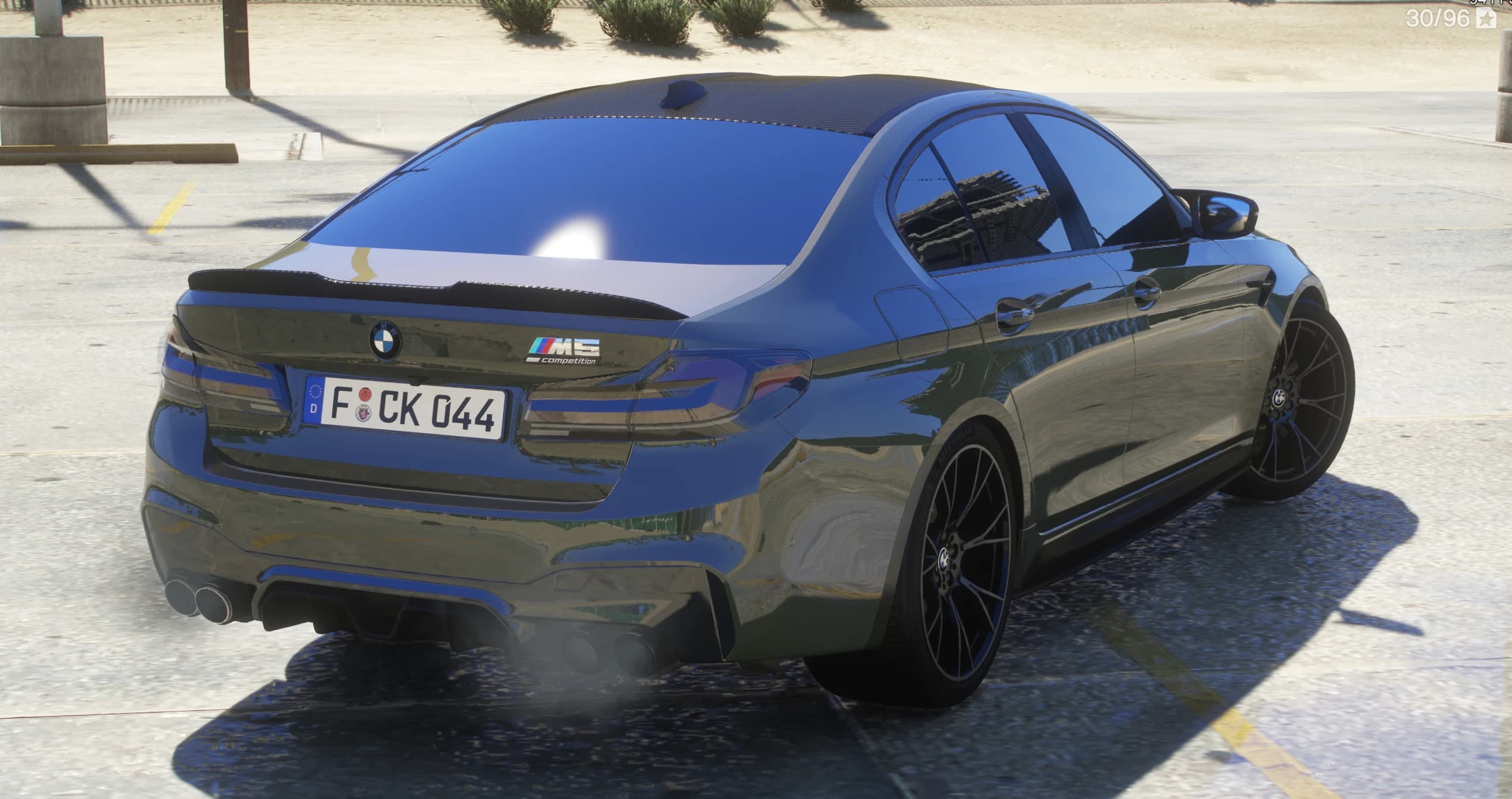Rear three-quarter of the olive M5 showing the carbon-fibre roof, ducktail spoiler with the M5 Competition badge, quad exhausts trailing smoke and black multi-spoke wheels on a sunlit lot