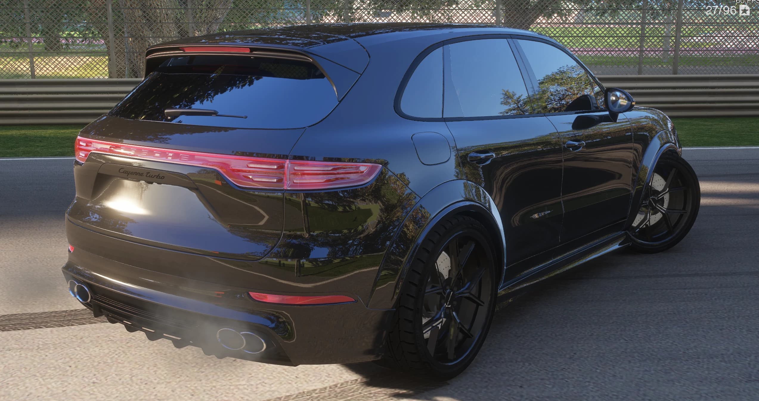 Rear three-quarter of the black Cayenne with the illuminated Porsche light bar lit across the tailgate, the Cayenne Turbo script, the roof spoiler and the exhausts blowing smoke next to the trackside barrier