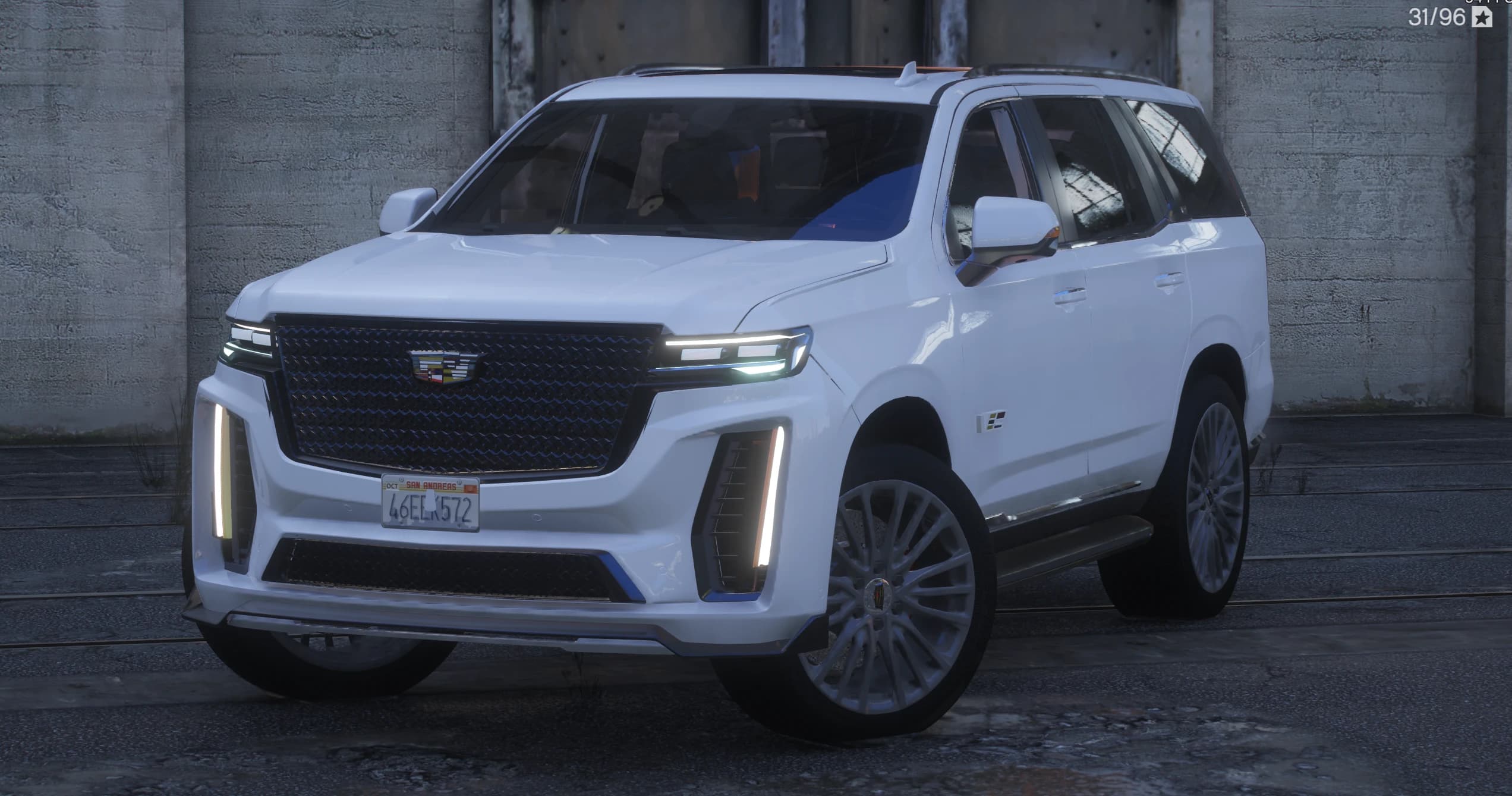 Front three quarter view of the white Escalade V with the black diamond mesh grille, vertical LED daytime lights and the V-Series badge on the door, parked by a concrete wall