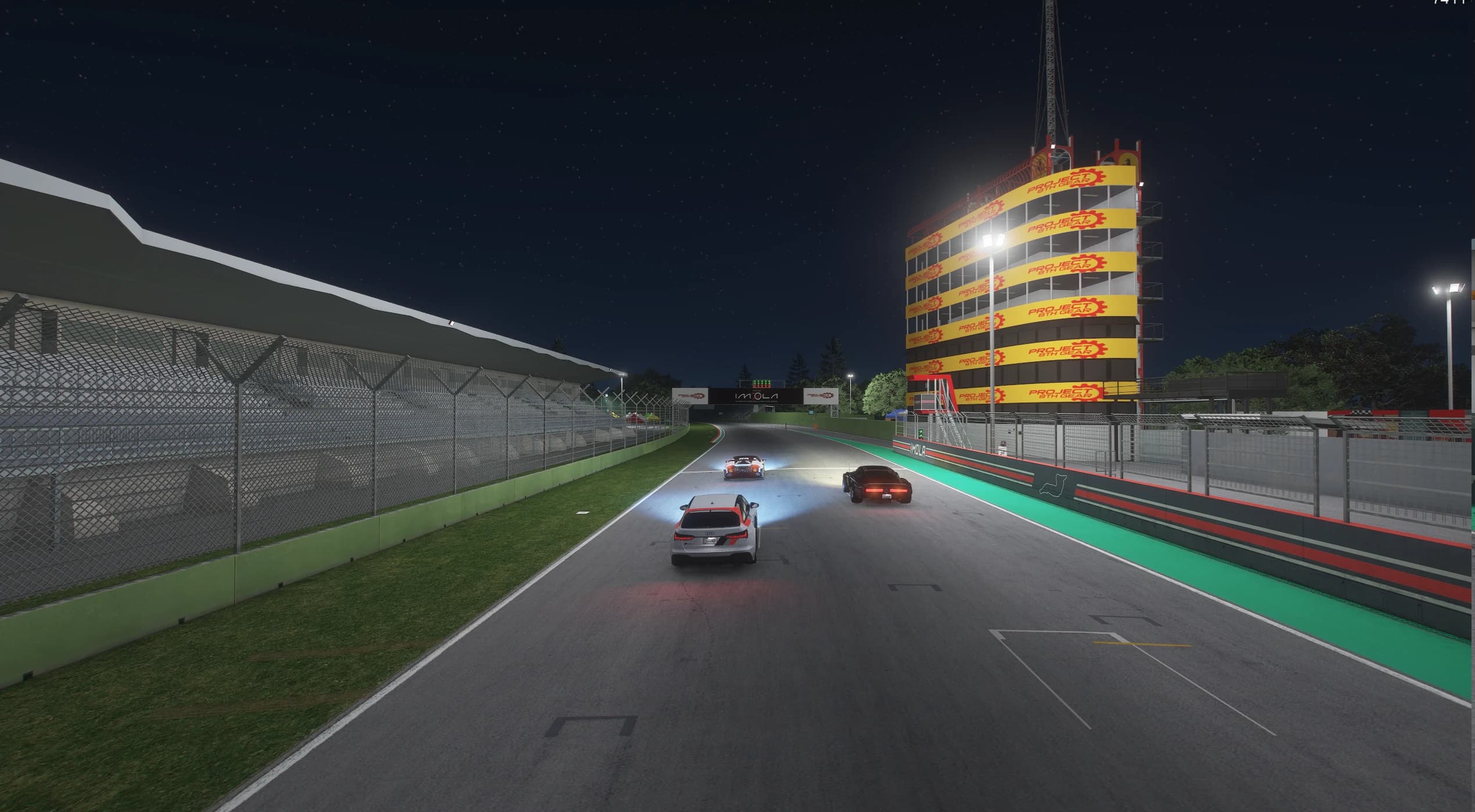 Three cars running down the floodlit pit straight at night, with the covered grandstand on the left and the tall pit tower covered in PROJECT 8TH GEAR banners on the right