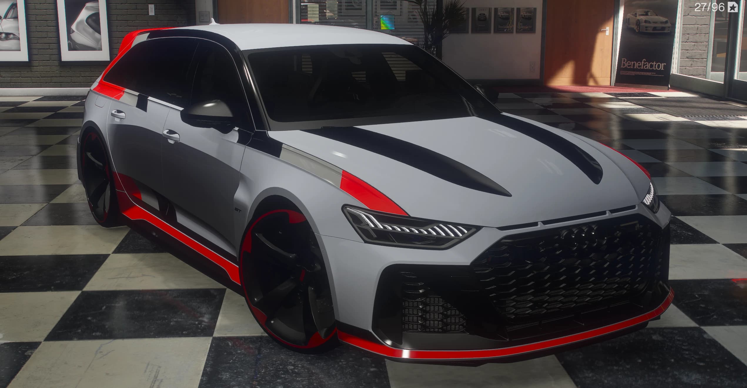 2025 Audi RS6 GT estate in nardo grey with red and black GT graphics, front three-quarter on the chequered floor of a car showroom, black honeycomb grille and black wheels with red rim edges