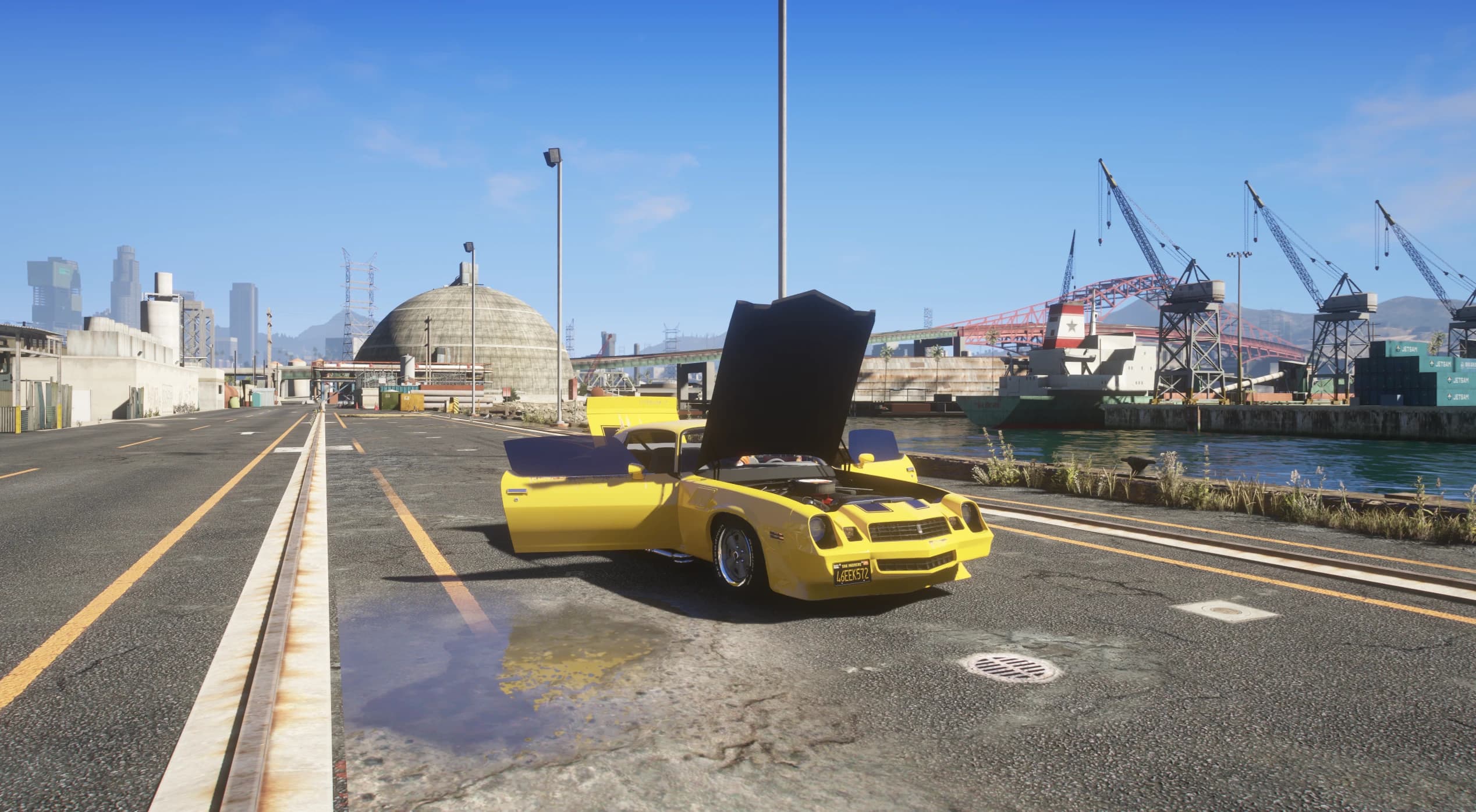 The Camaro with doors and hood open on a dock road, with the engine visible and cranes and a cargo ship behind