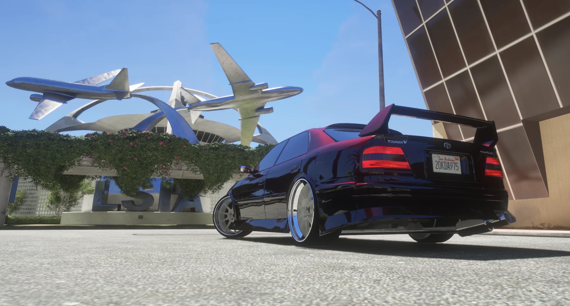 Rear view of the Chaser with its tall rear wing and Tourer V badge, parked in front of the LSIA airport sign and its jet sculpture