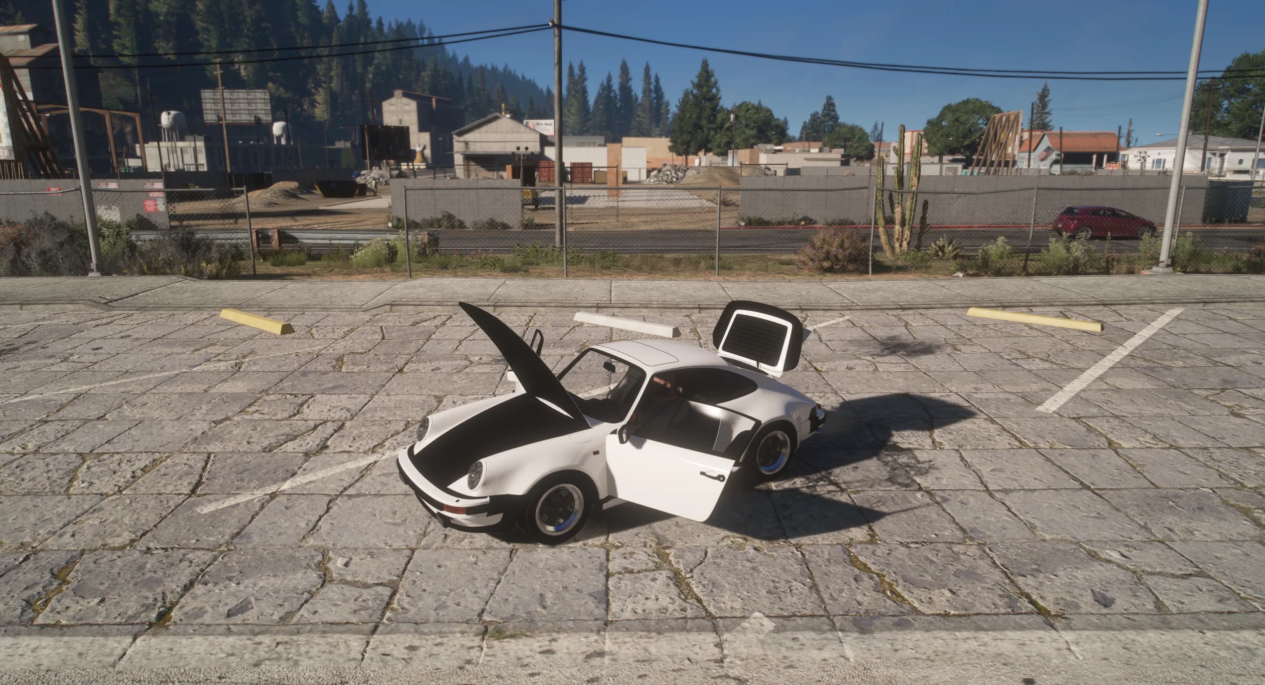 Side view of the white 911 Turbo with the front luggage lid, driver door and rear engine deck all open in a parking lot, with pine-covered hills and warehouses behind the chain-link fence