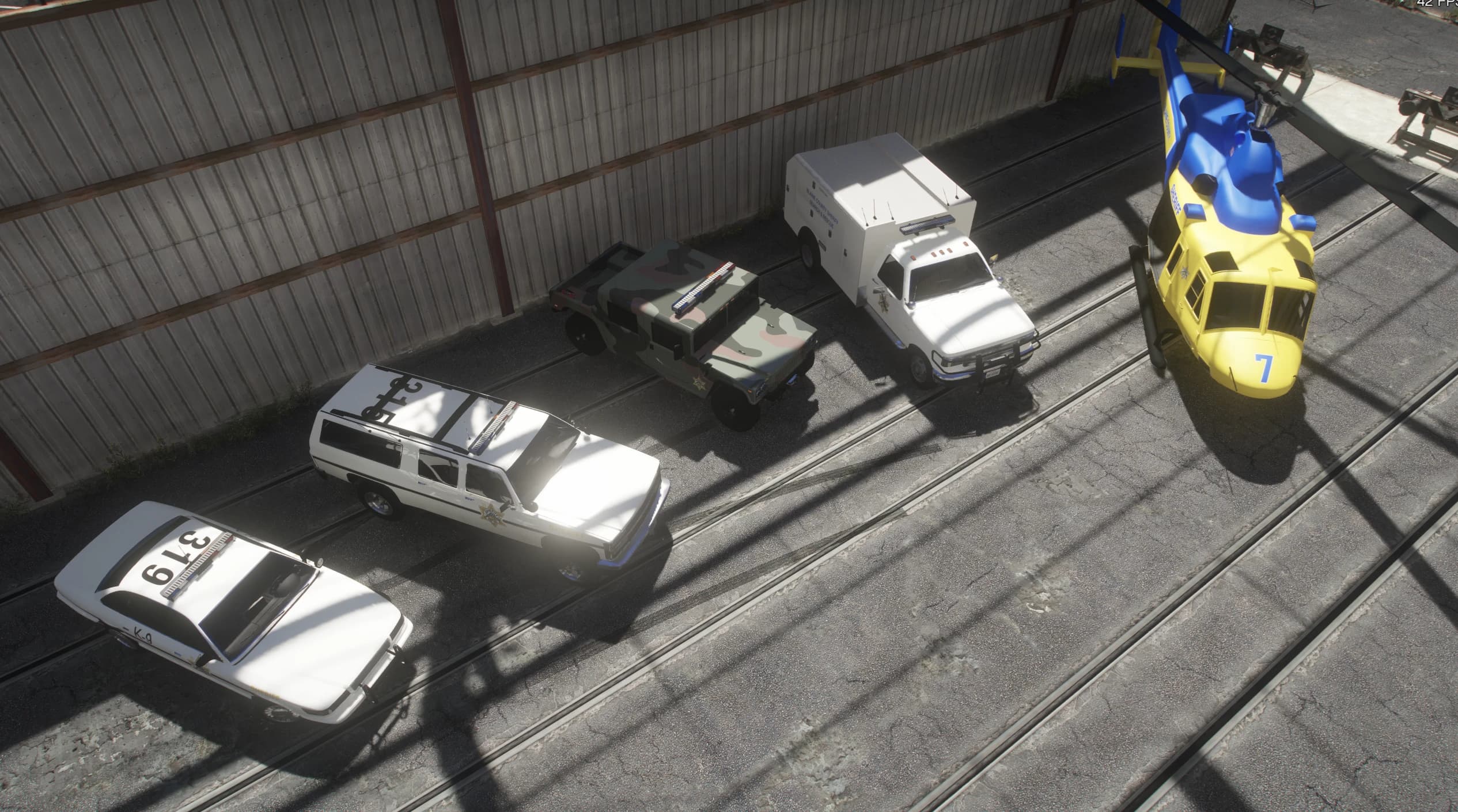 Aerial view inside a depot of a retro Blaine County Sheriff fleet: a white K-9 patrol car, a white sheriff SUV, a camo sheriff Humvee, a white sheriff rescue truck and a yellow-and-blue sheriff helicopter