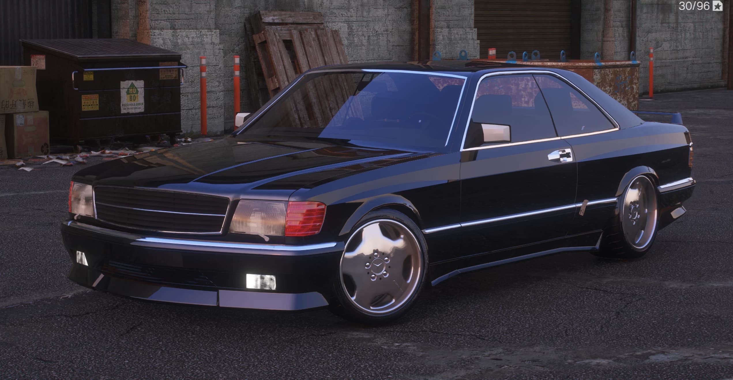 Black 1987 Mercedes-Benz 560 SEC coupe, front three-quarter view in an industrial alley beside a dumpster, showing the tall chrome grille with the three-pointed star, twin rectangular headlights and AMG-style chrome monoblock wheels