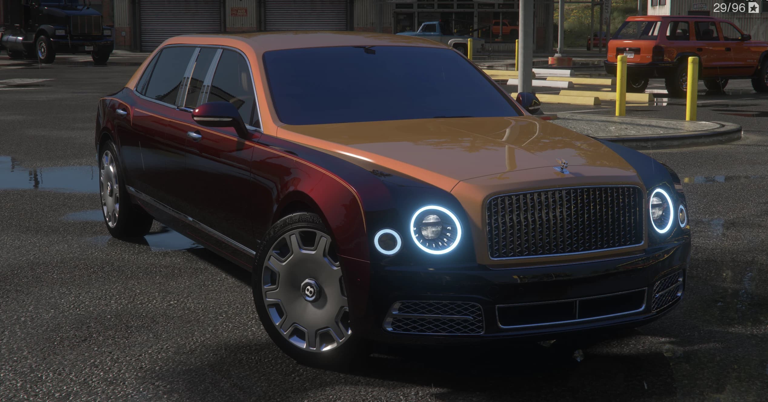 Two-tone Bentley Mulsanne limousine in maroon and copper, front three-quarter view with illuminated ring headlights, the winged Bentley badge on the grille and B-logo hubcaps, at a truck stop