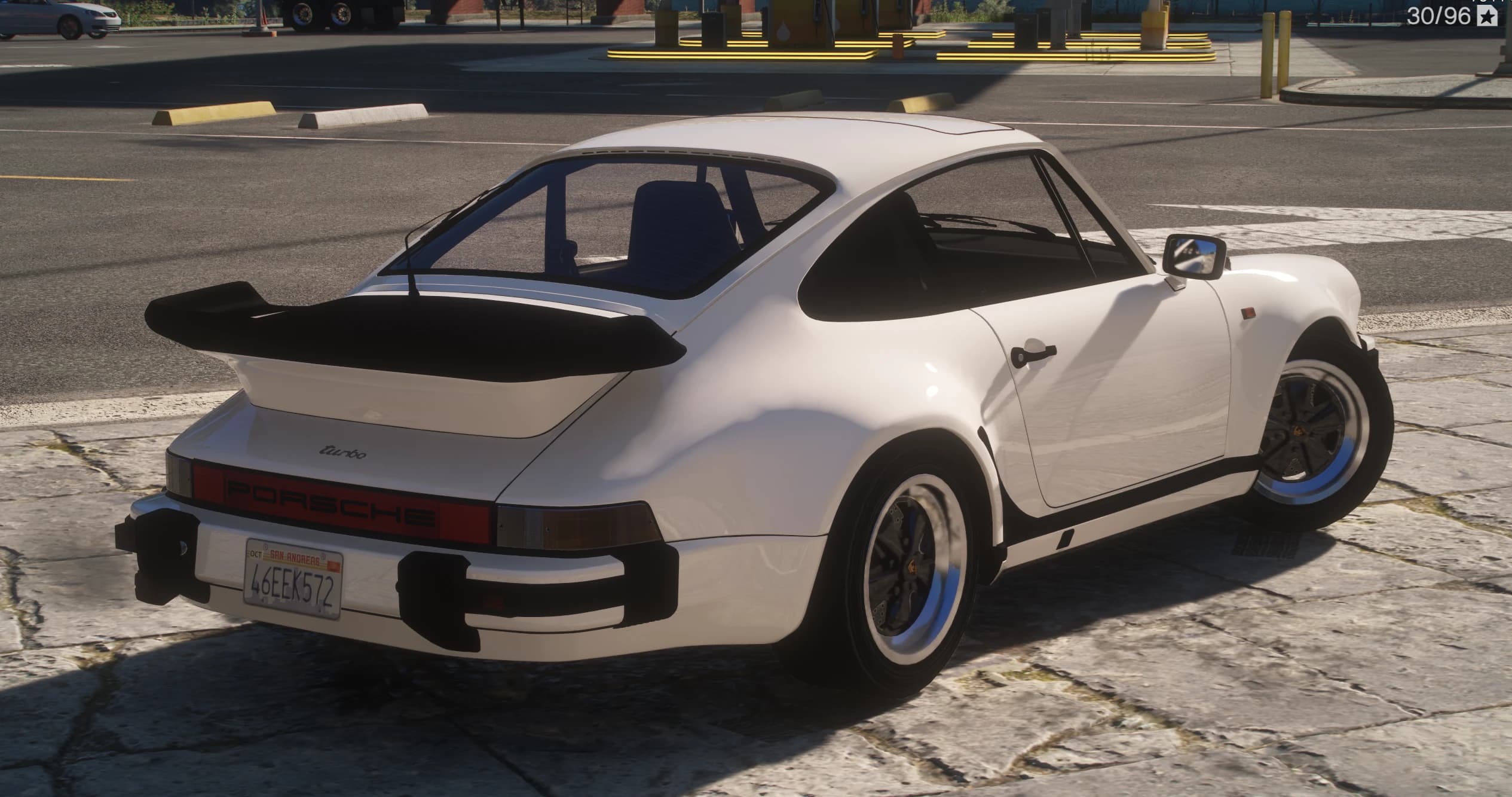 Rear three-quarter of the white 911 Turbo at a fuel station, showing the black-topped whale-tail spoiler, the 'turbo' script on the engine lid, the red 'PORSCHE' reflector band and a San Andreas plate reading 46EEK572