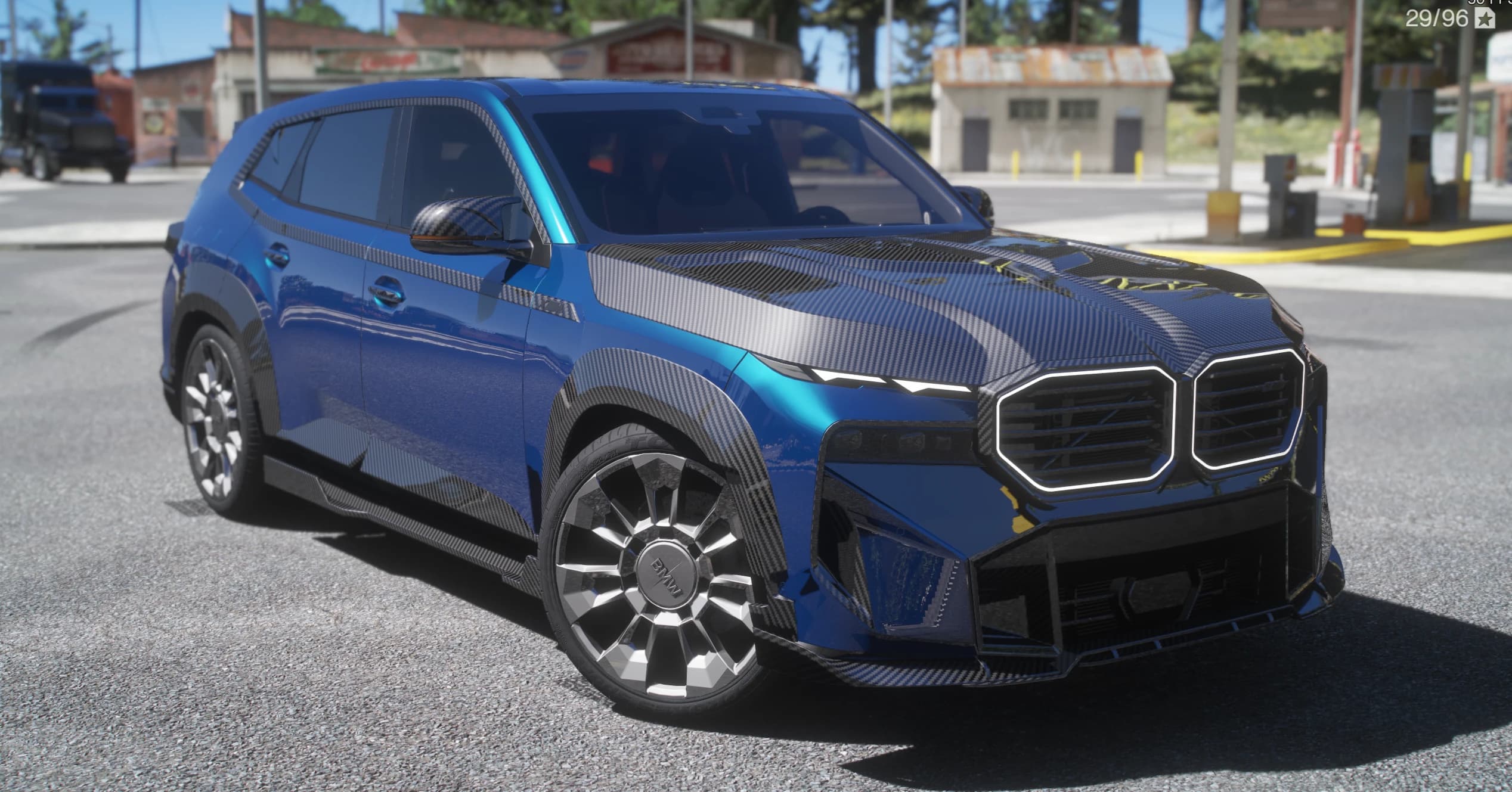Blue BMW XM G09 SUV with carbon panels and an illuminated kidney grille, front three-quarter view at a gas station