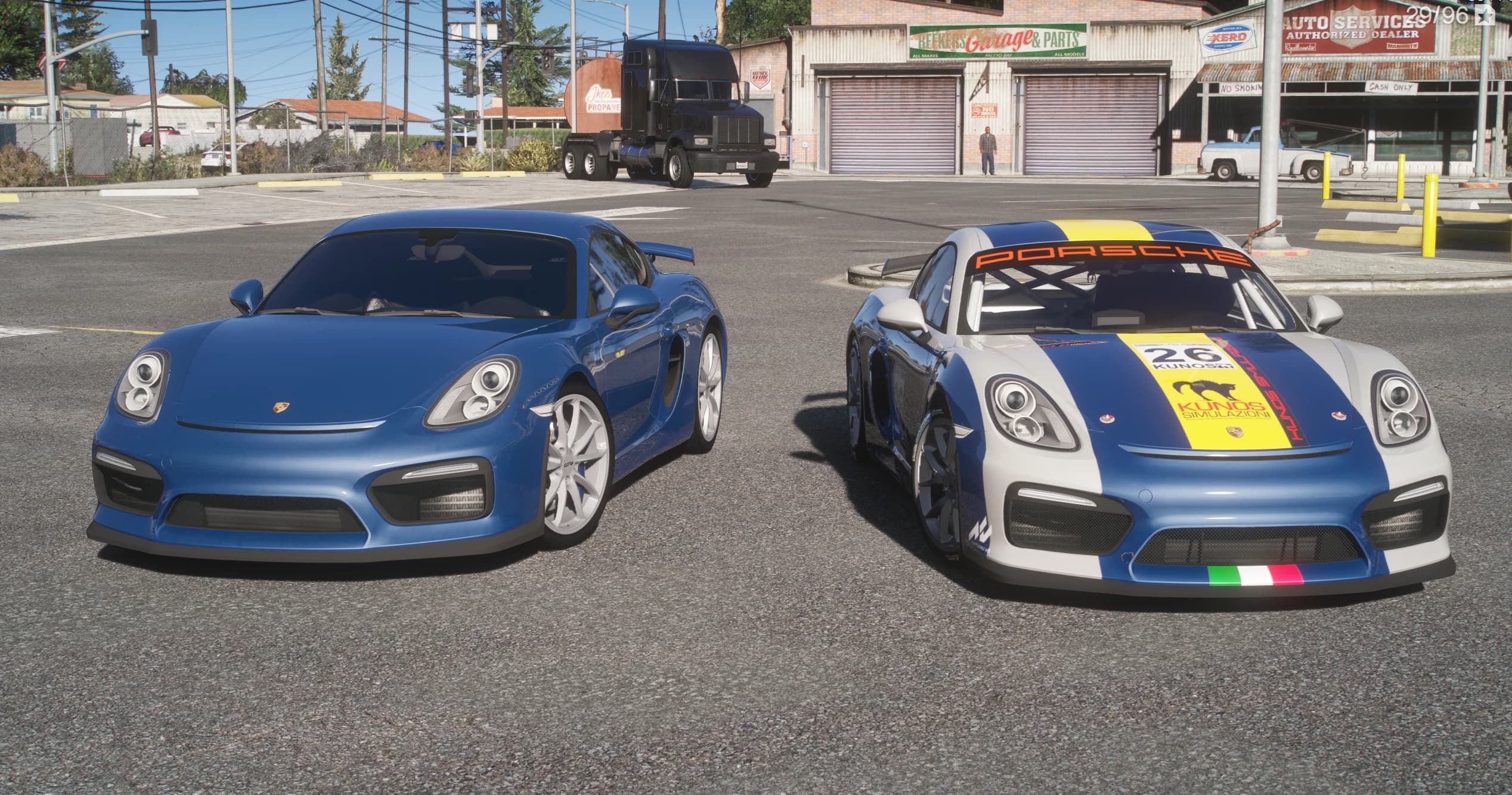 Blue road-going Porsche Cayman GT4 on the left beside the white-and-blue GT4 Clubsport race car with number 26 and a yellow stripe, parked outside an auto garage