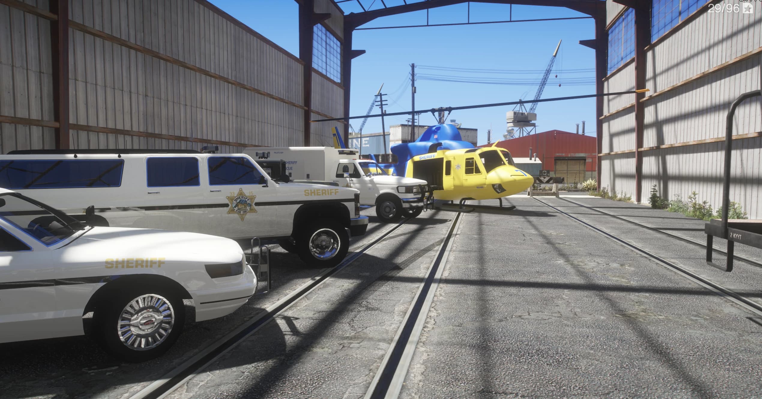 Ground-level view of the sheriff fleet lined up in a shed: a sheriff cruiser, a sheriff SUV, a Paleto Bay Sheriff Rescue truck and a yellow sheriff helicopter, all in the retro livery