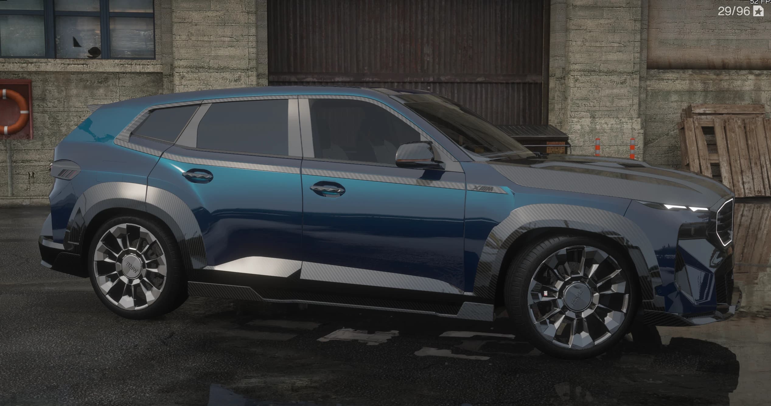 Side profile of the blue BMW XM with a carbon lower-body wrap, XM badge and large multi-spoke wheels beside an industrial building