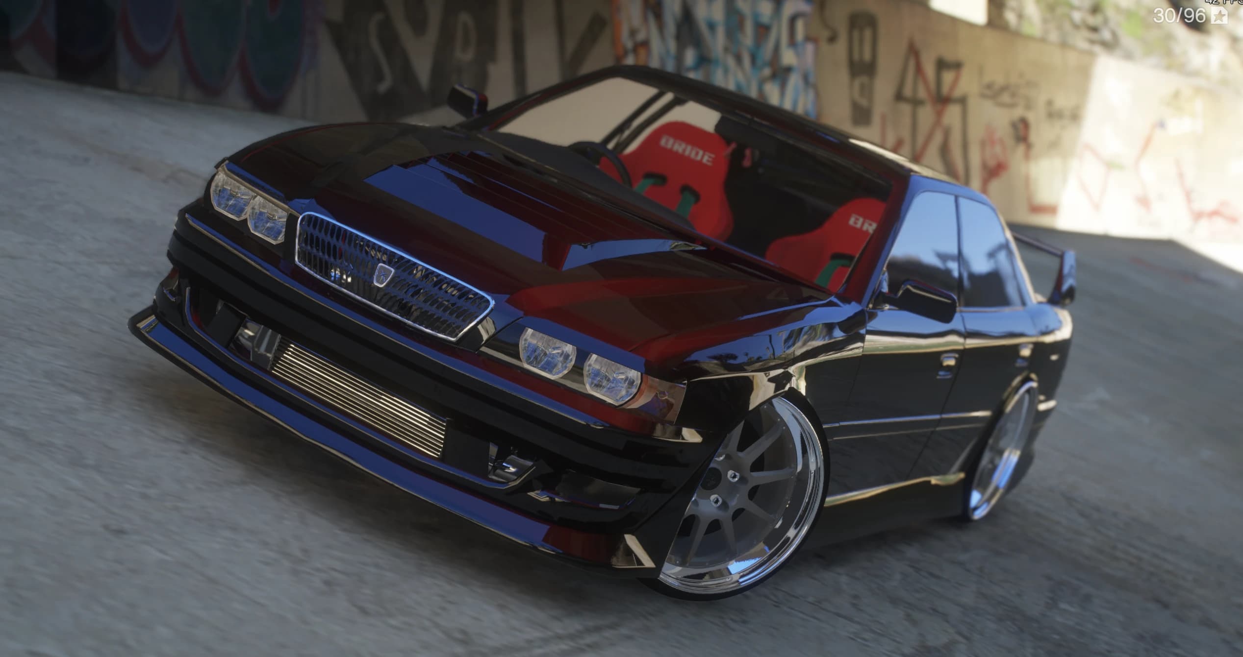 Front three quarter view of the black Toyota Chaser JZX100 under a graffiti-covered underpass, showing the exposed front-mount intercooler, quad headlights and red BRIDE-style bucket seats behind the windscreen