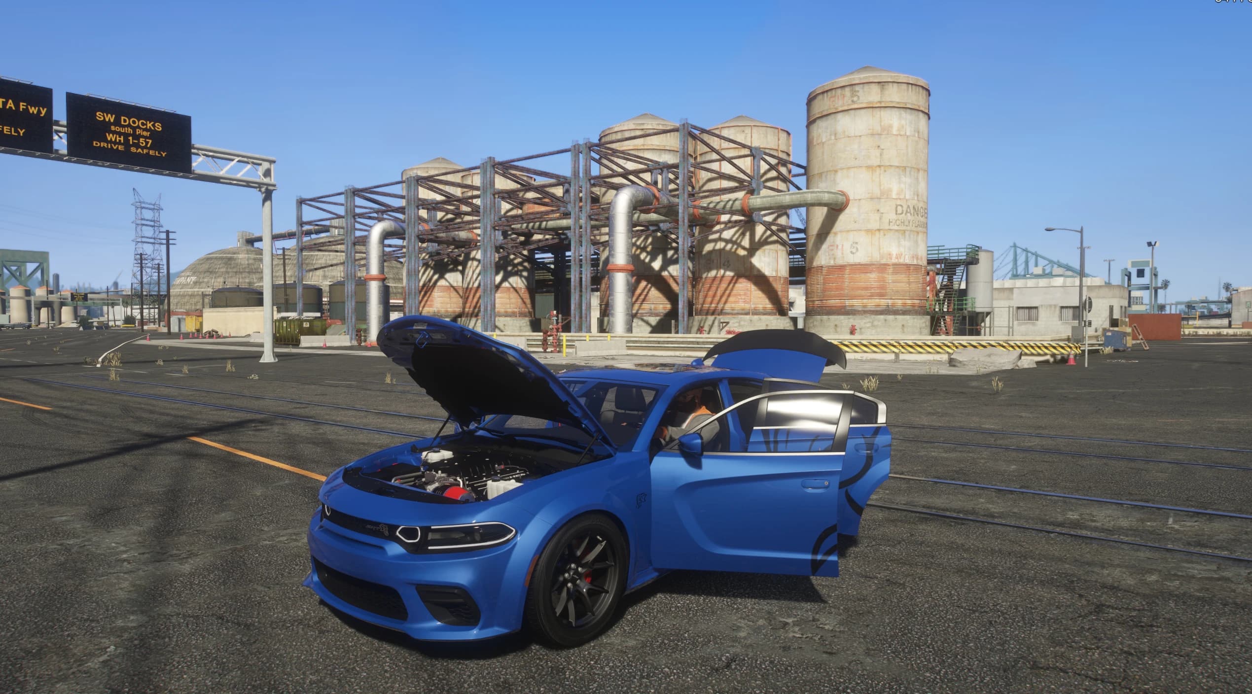 Blue Charger with the hood and driver door open at the docks, revealing the supercharged HEMI engine bay with a red intake, beneath a 'SW DOCKS' gantry sign and large flammable-storage tanks