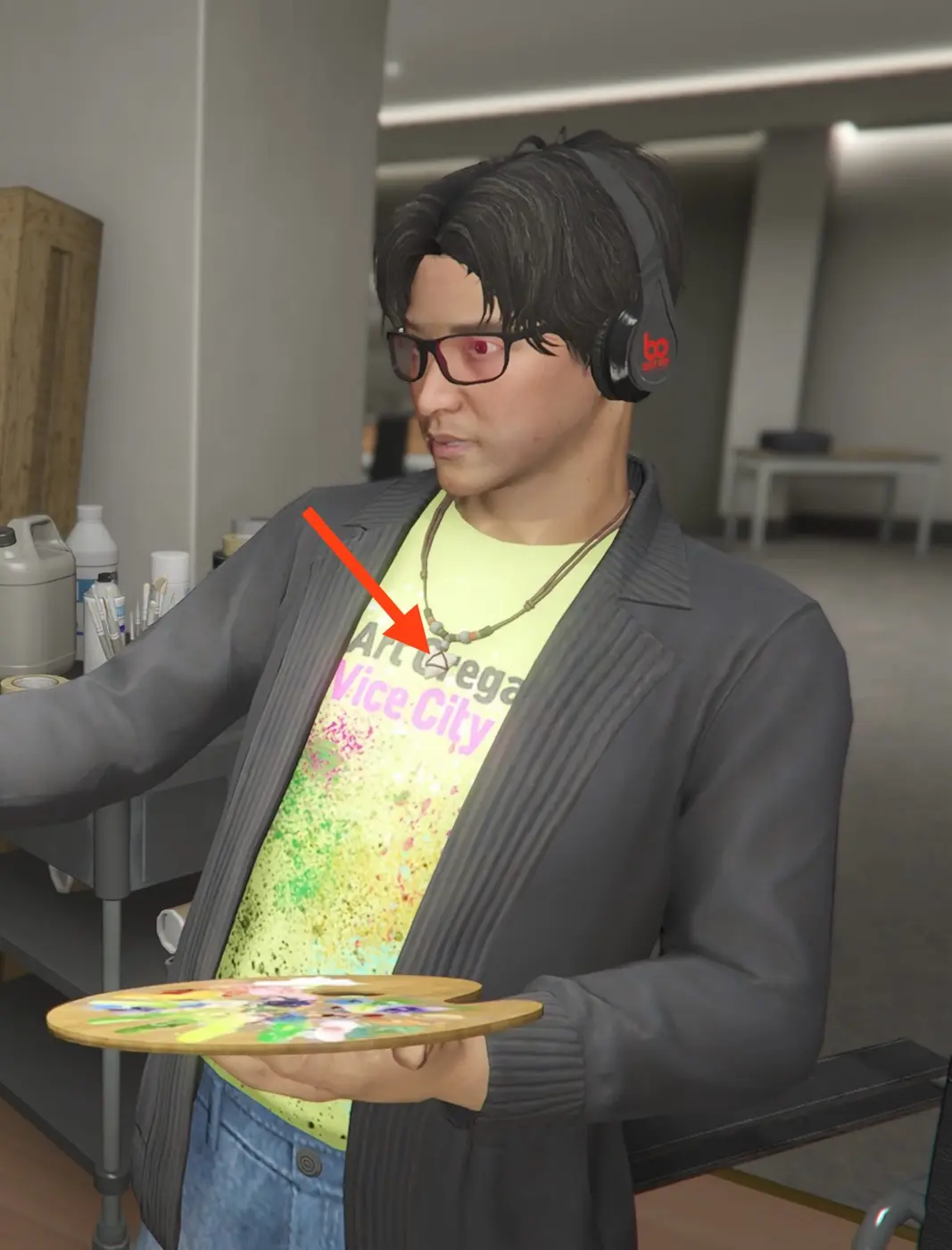 A GTA Online character with glasses and headphones holding a painter's palette, wearing a bright yellow t-shirt that reads 'Art Gallery Vice City' in pink lettering, with a red arrow pointing at an hourglass pendant on a beaded necklace.